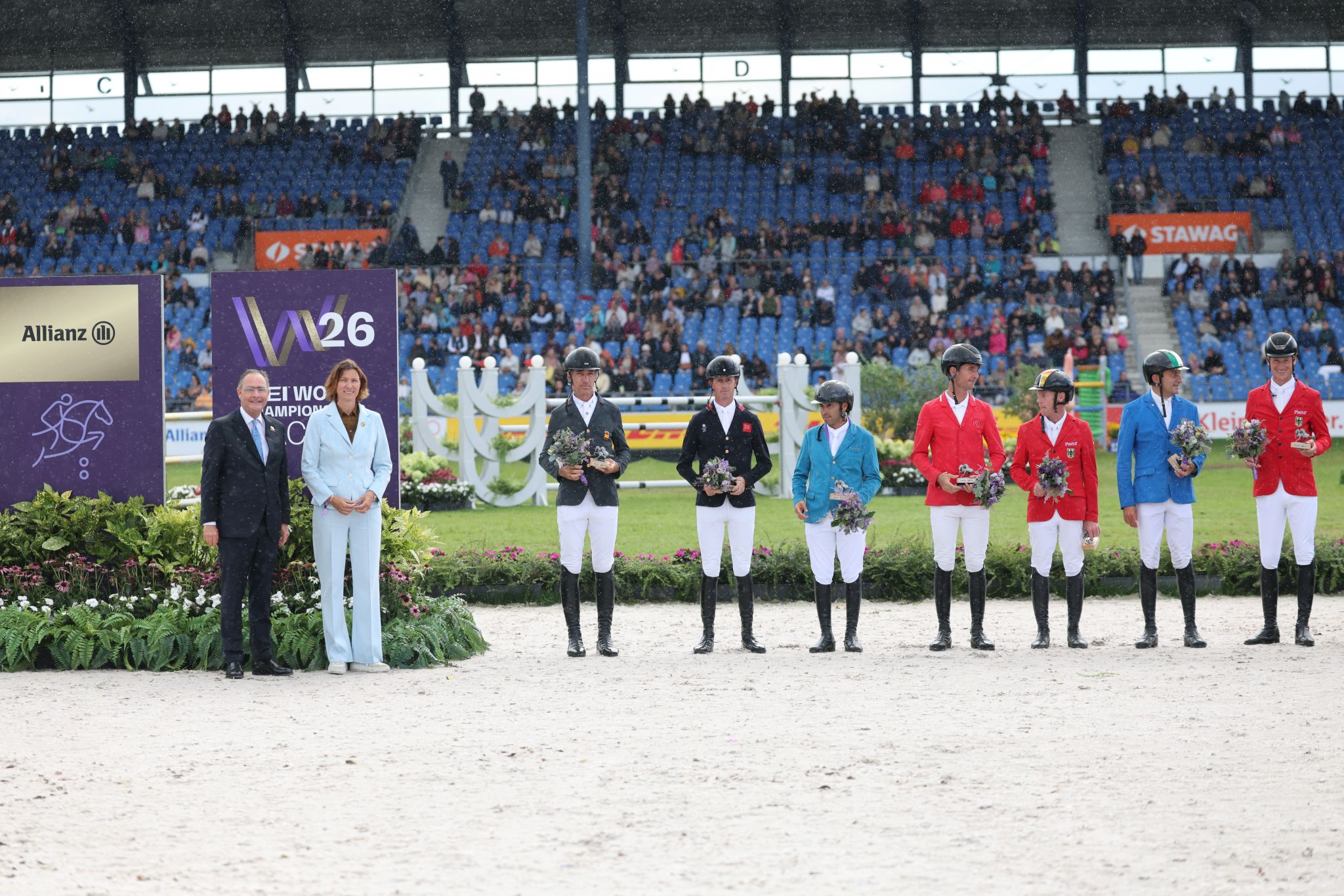 Prize-giving ceremony for jumping: Six riders wearing different-colored competition jackets stand with flowers in the Aachen Main Stadium. Two guests of honor stand next to them; in the background, spectators sit in the stands.