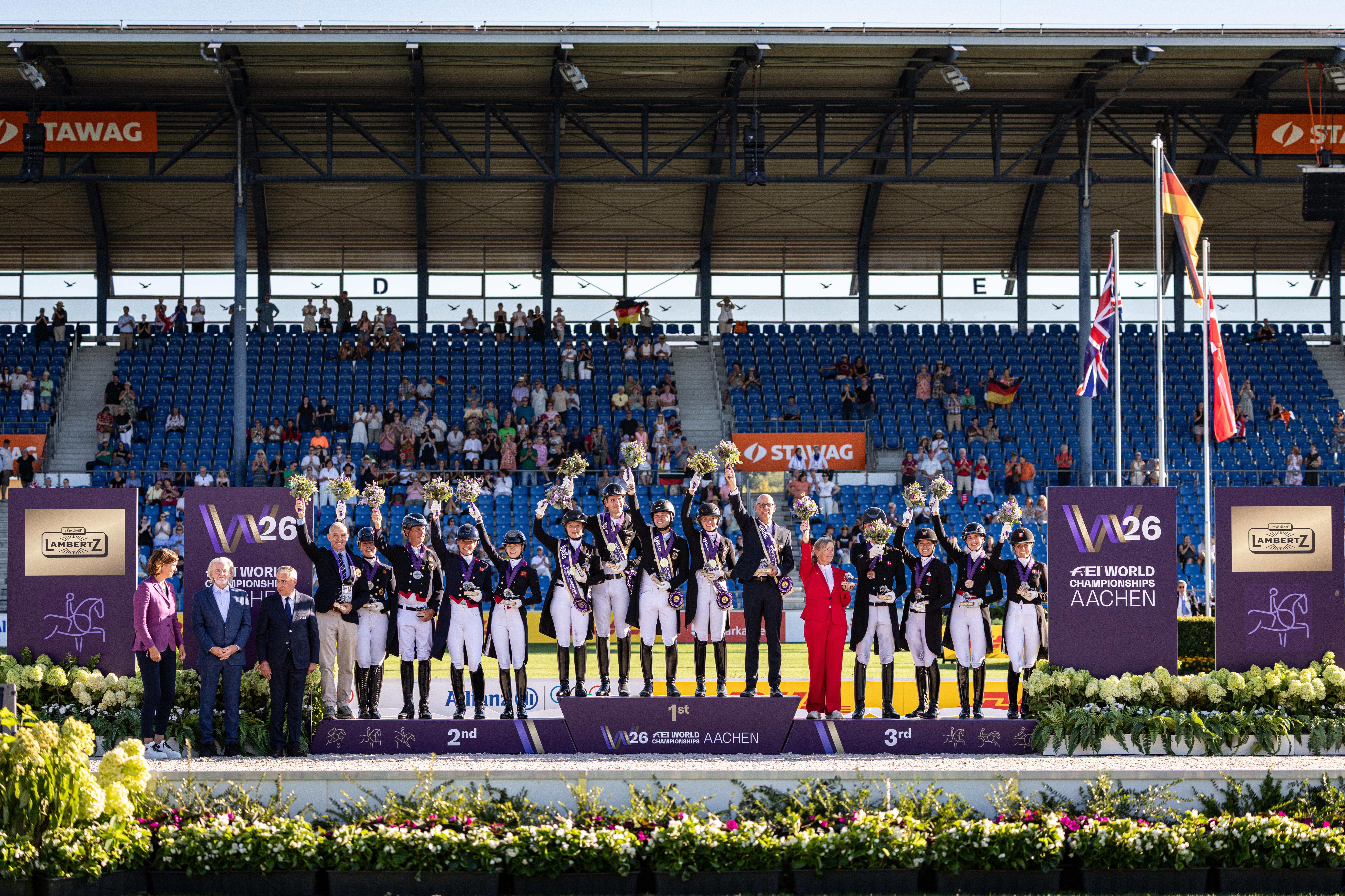 Siegerehrung im Reitstadion Aachen: Drei Dressurmannschaften stehen in Reituniformen auf dem Siegerpodest der FEI World Championships Aachen, heben ihre Blumensträuße in die Höhe und tragen Medaillen. Im Hintergrund sind Tribünen mit Zuschauern sowie Fahnen mehrerer Nationen zu sehen, links und rechts rahmen violette Lambertz- und W26 Branding-Wände die Szene ein.