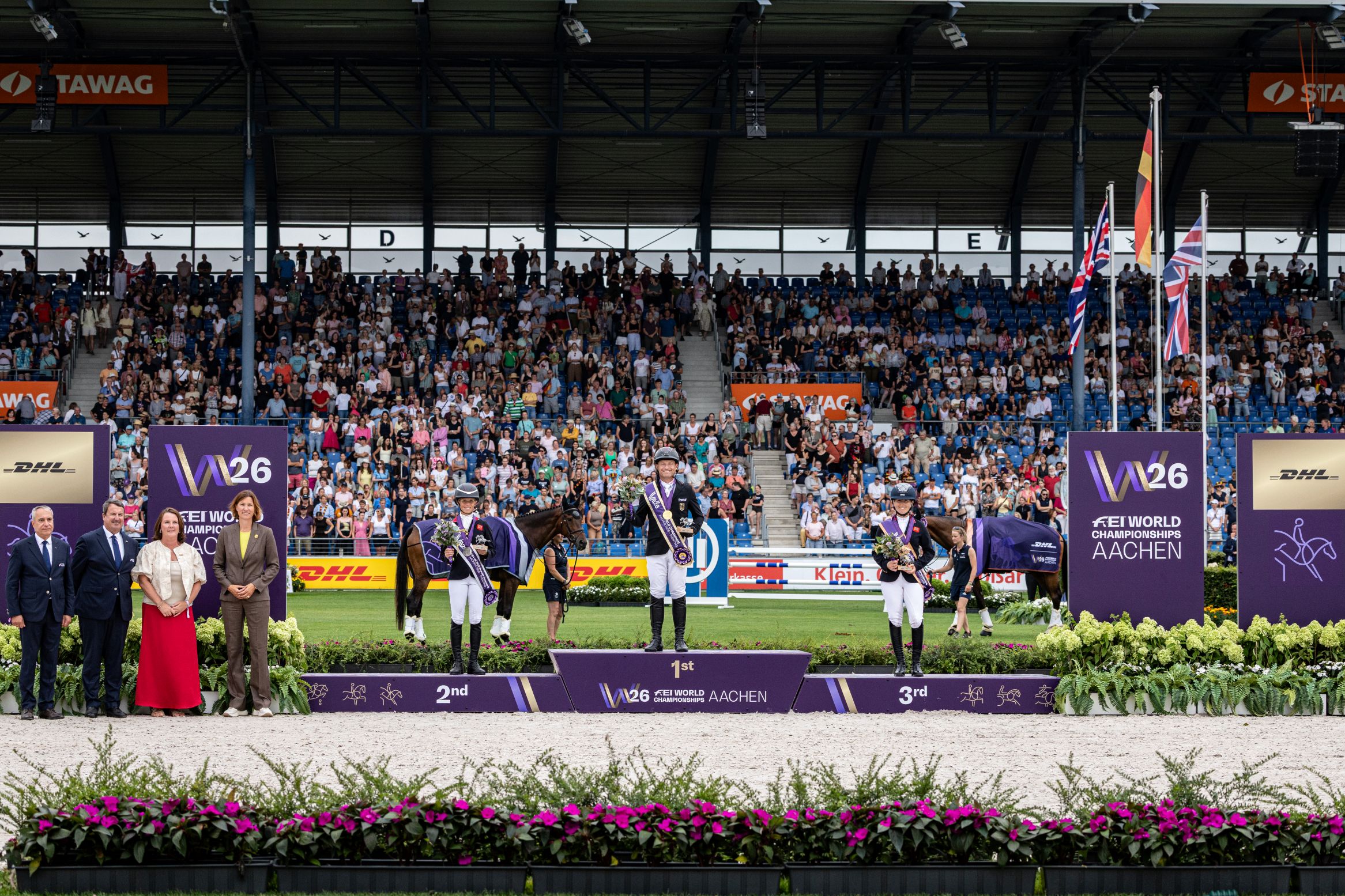 Medal Ceremony for the individual eventing competition at the Aachen Main Stadium: Three riders in competition attire stand on the winners’ podium with their decorated horses, with the inscriptions ‘1st’, ‘2nd’ and ‘3rd’ in front of packed stands, flanked by officials on the left and purple W26 and DHL branding boards and fluttering national flags on the right; in the foreground, flower beds line the sand arena.