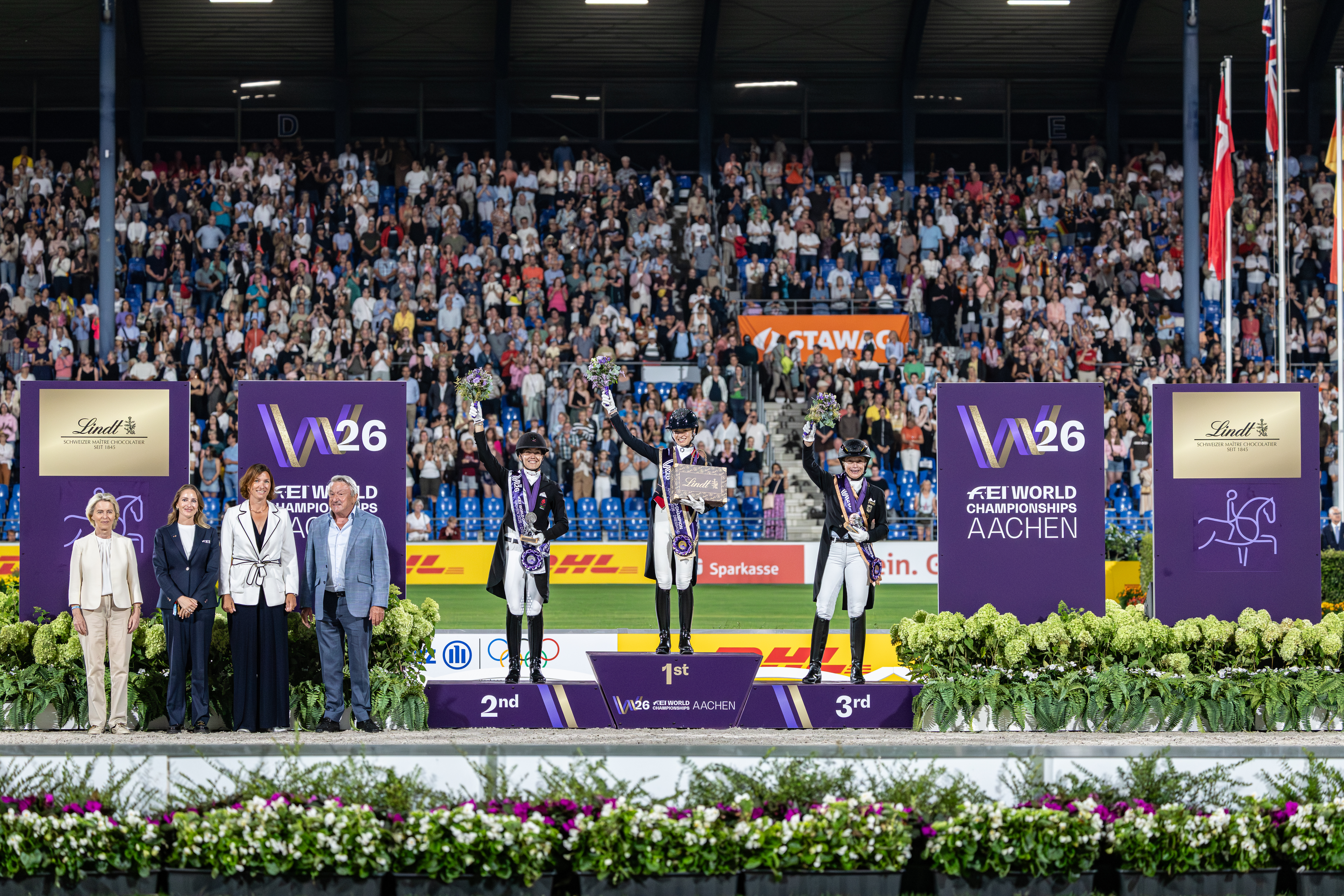 Siegerehrung Dressur bei den FEI World Championships Aachen 2026: Drei Reiterinnen in Frack, Reithelmen und Schärpen stehen mit Blumensträußen jubelnd auf dem Podest vor einer voll besetzten, applaudierenden Tribüne, flankiert von Offiziellen und großen violetten Turnierwänden mit W26- und FEI-Branding.
