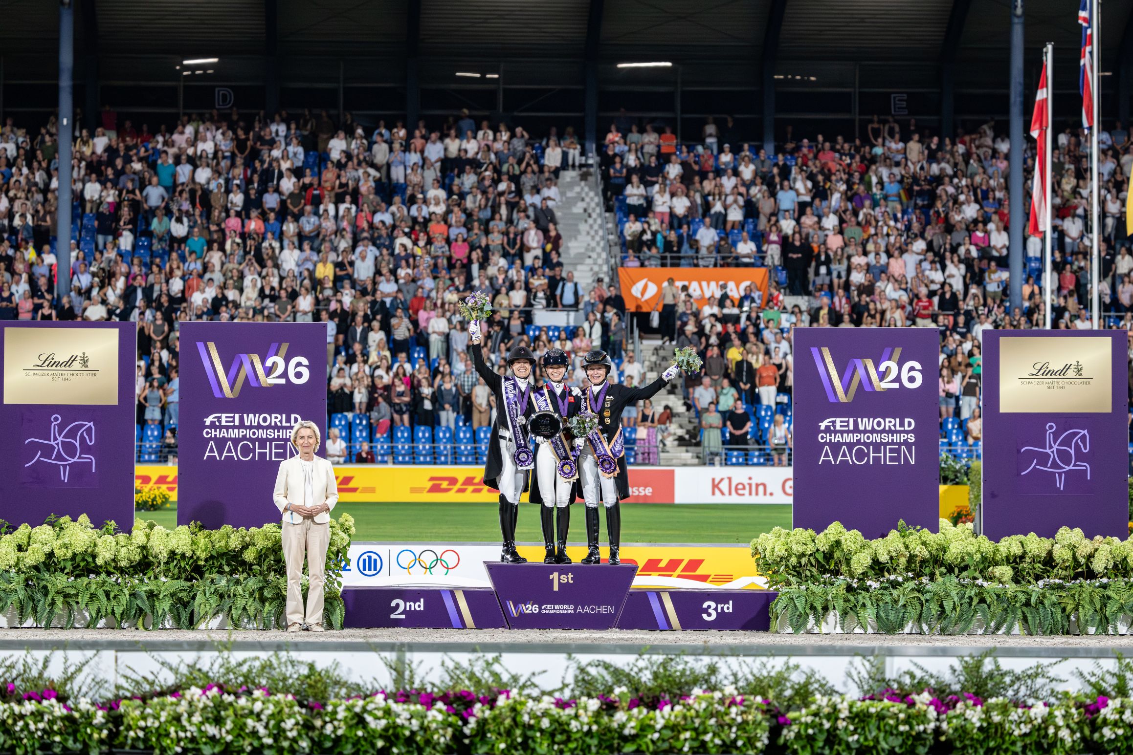 Prize-giving ceremony in a packed stadium: three female riders in competition attire stand on the podium, holding bouquets of flowers and wearing purple ‘W26 FEI World Championships Aachen’ sashes; to the left stands a woman in a light-coloured suit, flanked by large W26 and Lindt signs.
