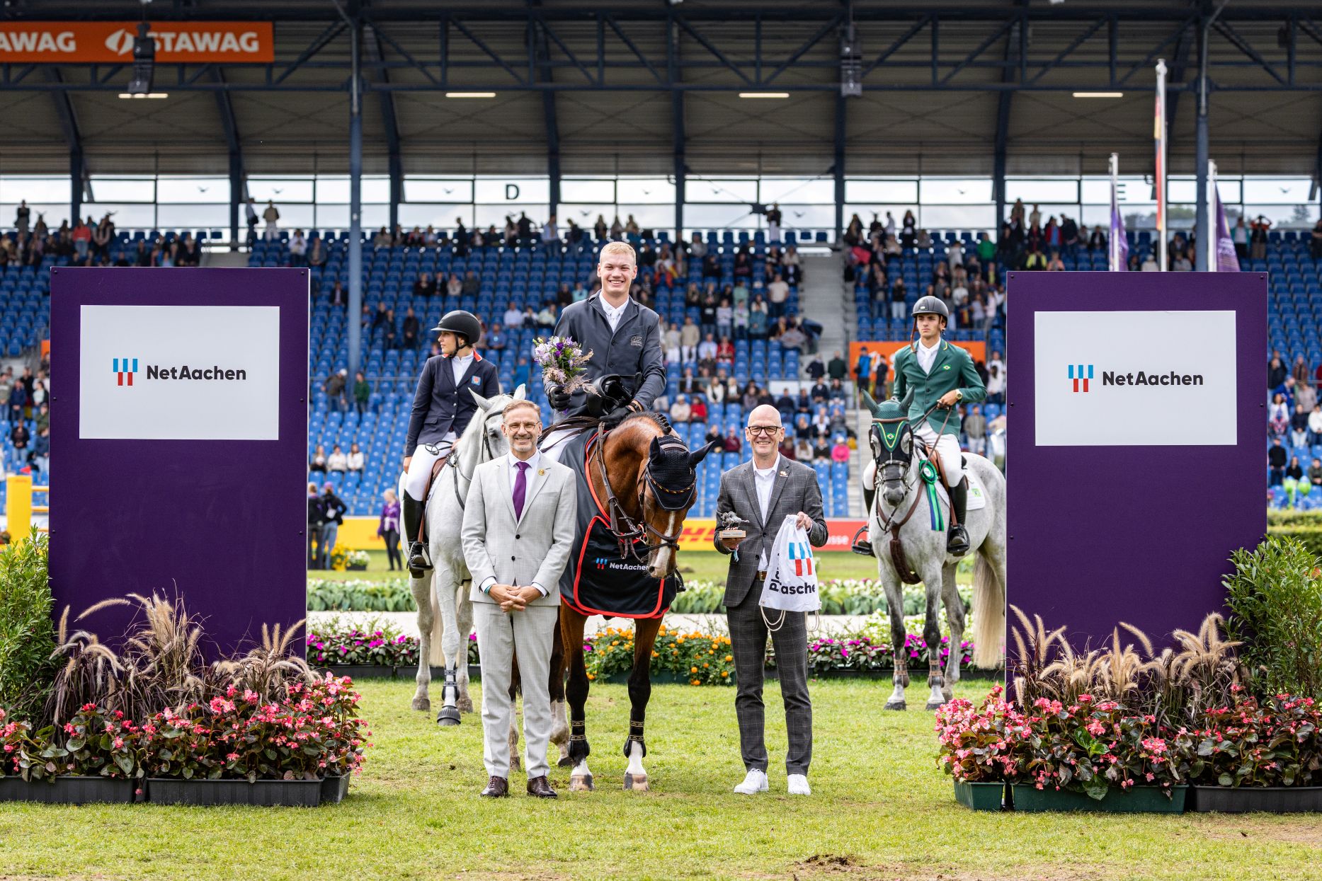 Prize-giving ceremony at an equestrian event in the stadium: a rider holding a bouquet of flowers sits on a bay horse, flanked by two other riders on white horses. Two men stand in front of them; in the background, spectators watch the ceremony. NetAachen advertising hoardings can be seen at the sides.