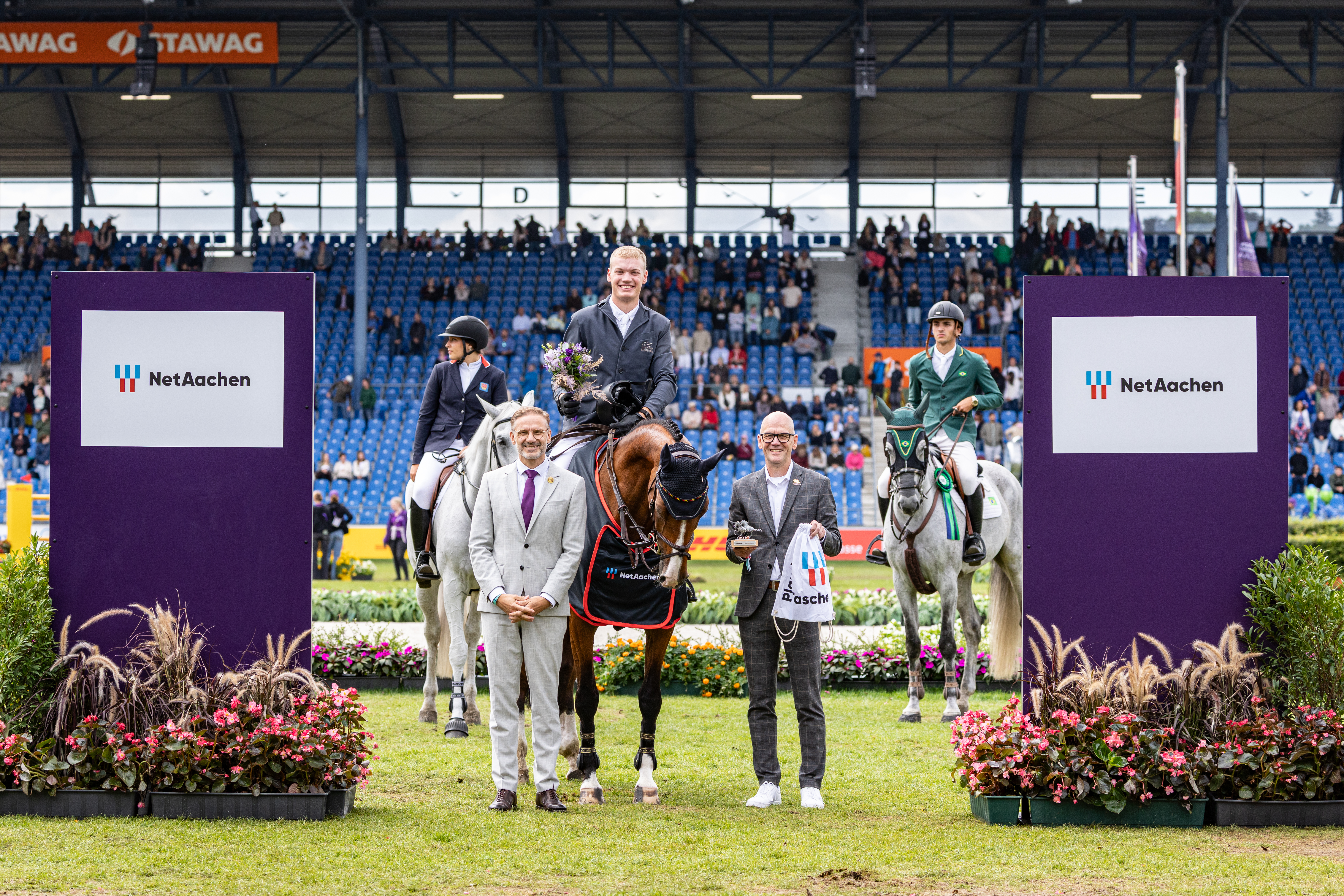 Siegerehrung bei einem Reitturnier im Stadion: Ein Reiter mit Blumenstrauß sitzt auf einem braunen Pferd, flankiert von zwei weiteren Reitern auf weißen Pferden. Zwei Männer stehen davor; im Hintergrund verfolgen Zuschauer die Zeremonie. Seitlich sind NetAachen-Werbetafeln zu sehen.