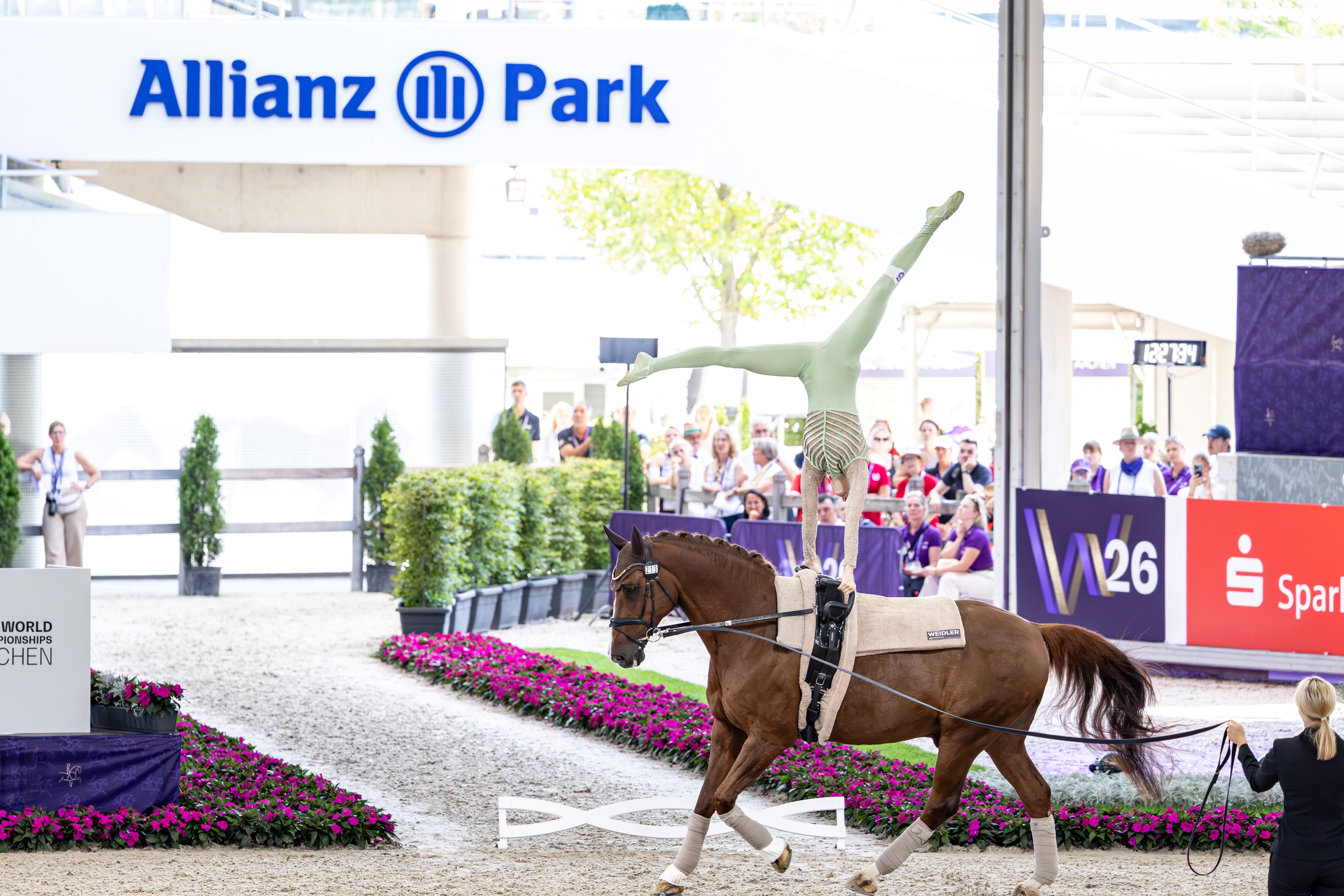 A vaulting rider in a green leotard performs a split whilst in a handstand on the back of a bay horse in the arena, led on the lunge line, with rows of spectators, flower beds and the ‘Allianz Park’ sign in the background.