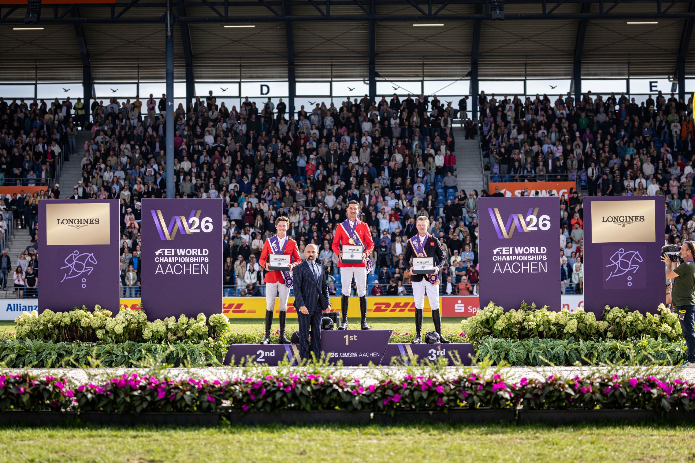 Three riders stand on the podium at the Medal Ceremony for the 2026 FEI World Championships in Aachen: the winner in the centre in first place, flanked by the runner-up and the third-placed rider. Behind them, the audience applauds; flowers, sponsors’ logos and signs bearing the words ‘FEI World Championships Aachen’ frame the scene.