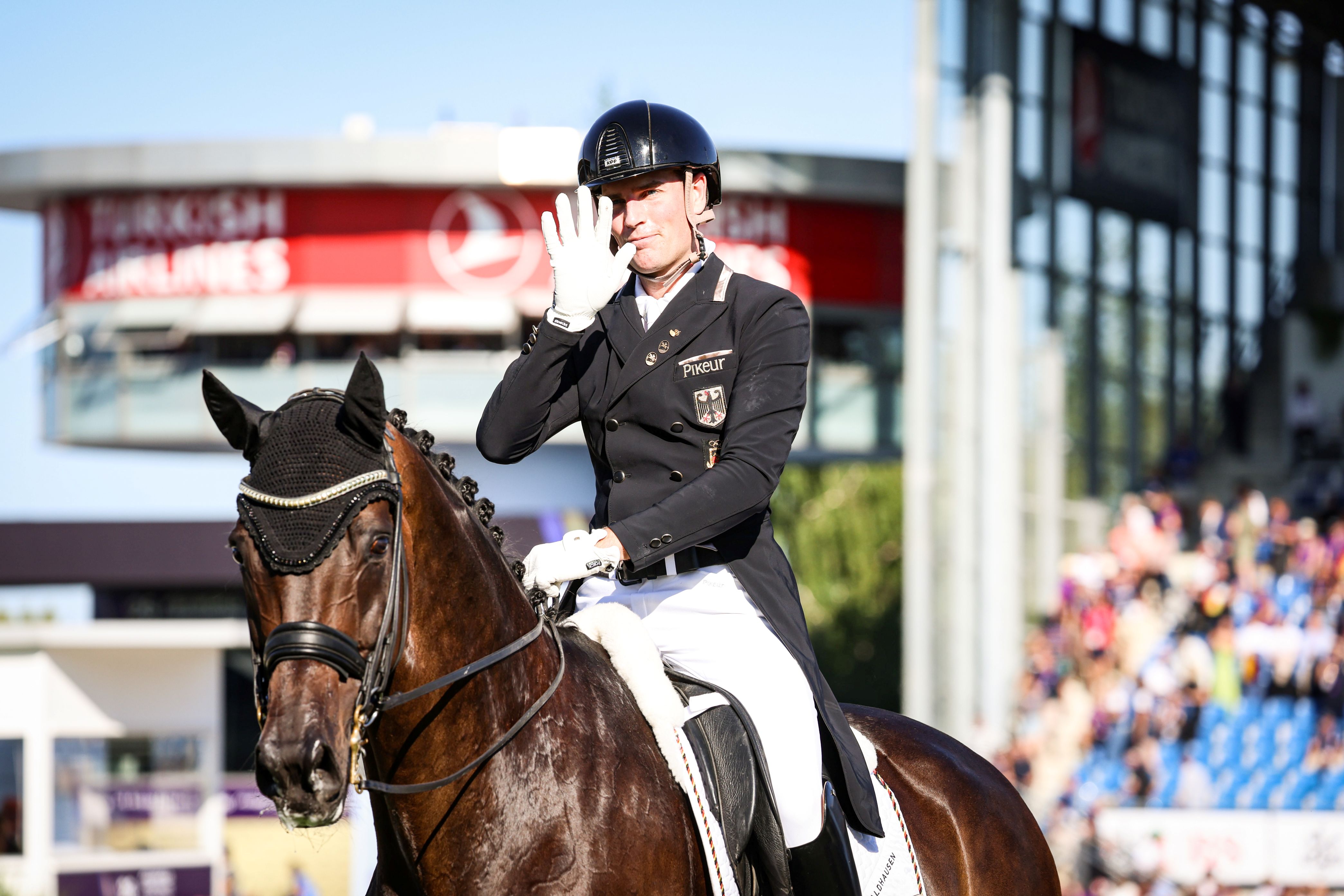 A dressage rider in a dark tailcoat, white breeches and black helmet raises a gloved hand in greeting while sitting on a dark bay horse with a braided mane and ear bonnet, in a sunny stadium with spectators and sponsor signage blurred in the background.