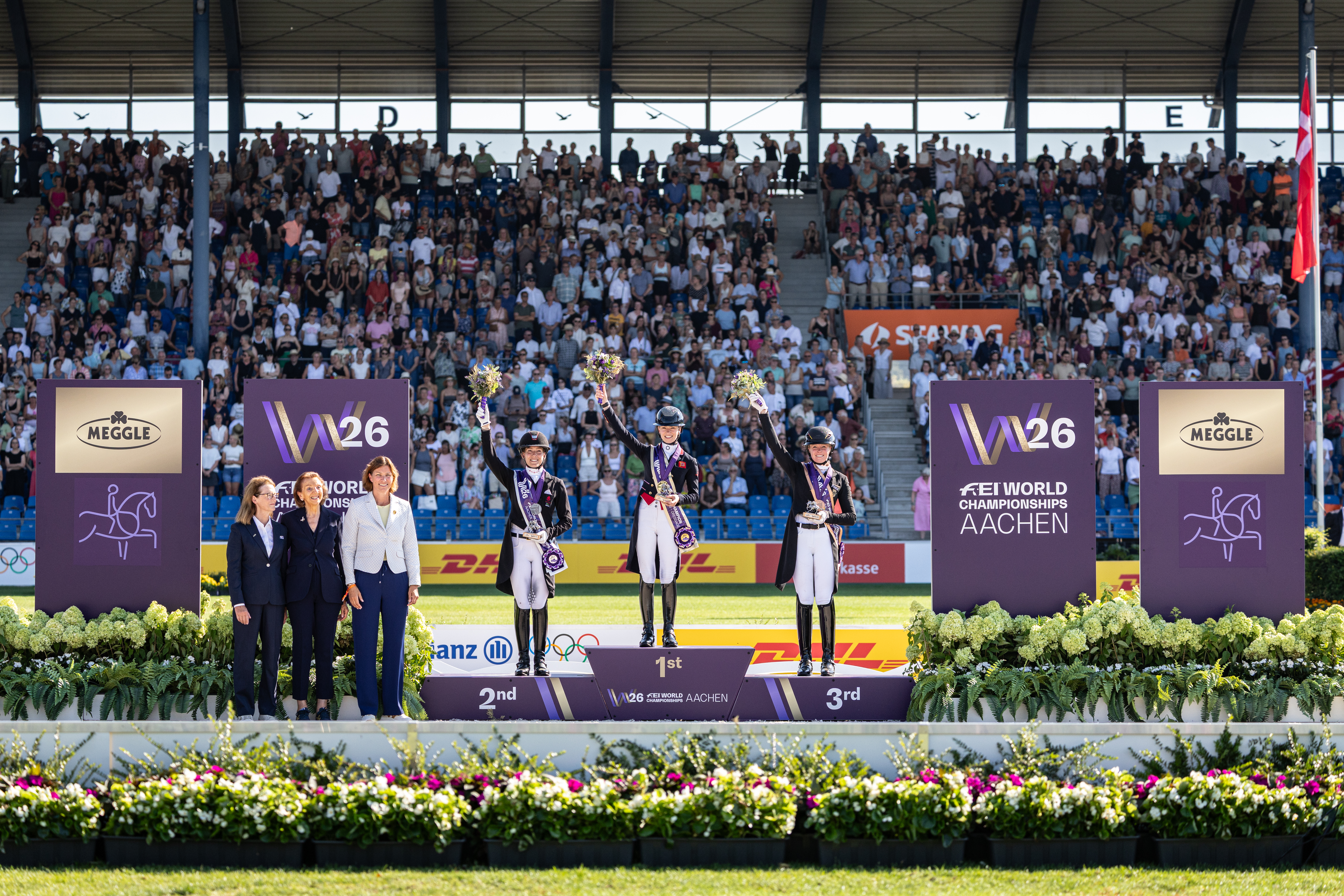 Siegerehrung im vollbesetzten Hauptstadion in Aachen: Drei Reiterinnen in Frack und Zylinder stehen auf dem Podium der FEI World Championships Aachen 2026, heben ihre Blumensträuße in die Höhe und tragen Medaillen, flankiert von Offiziellen vor lila Turnier- und Sponsorentafeln sowie üppigen Blumenarrangements.