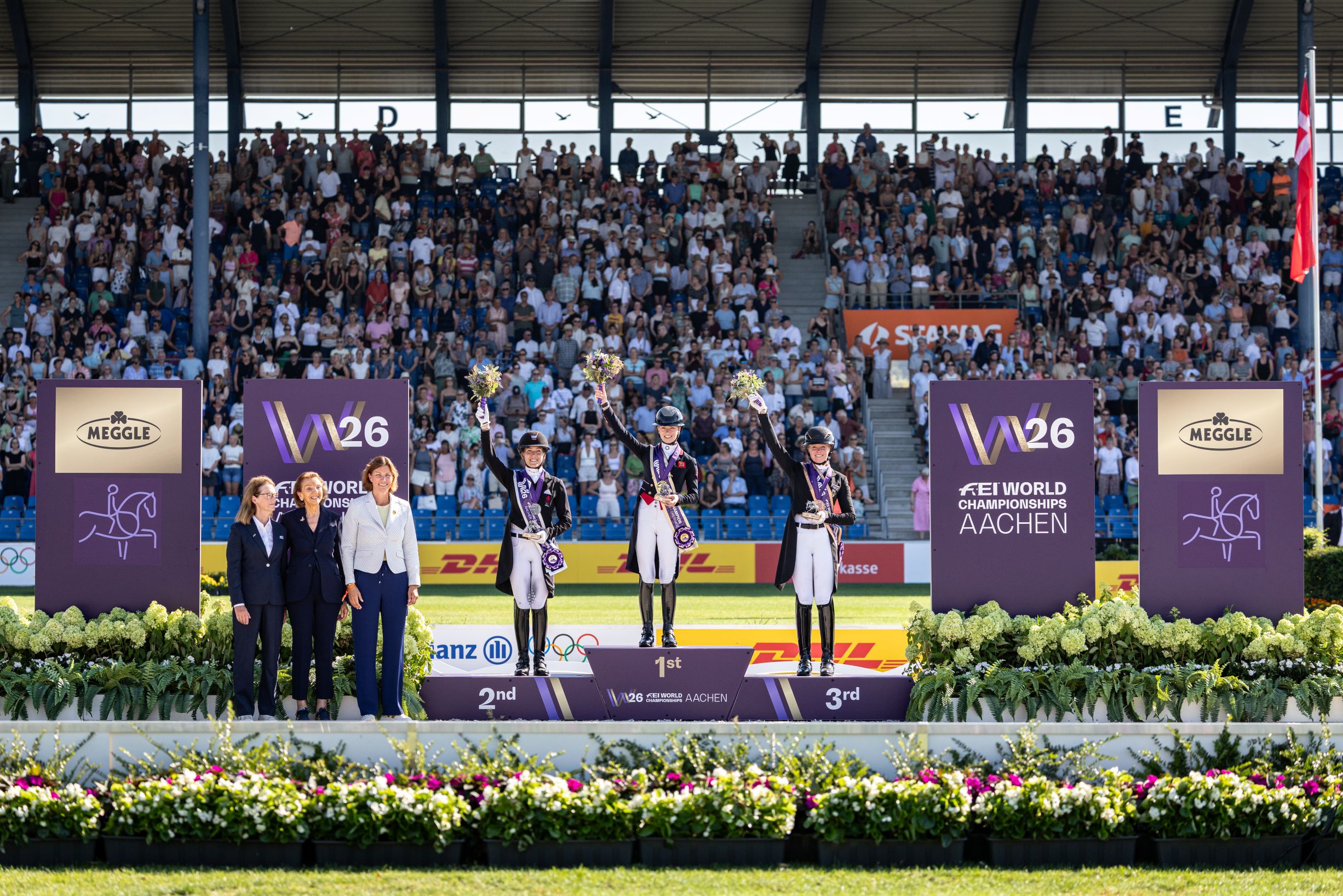 Medal ceremony in the packed Main Stadium in Aachen: three female riders in tailcoats and top hats stand on the podium at the 2026 FEI World Championships in Aachen, holding their bouquets aloft and wearing their medals, flanked by officials in front of purple tournament and sponsor boards and lavish floral arrangements.
