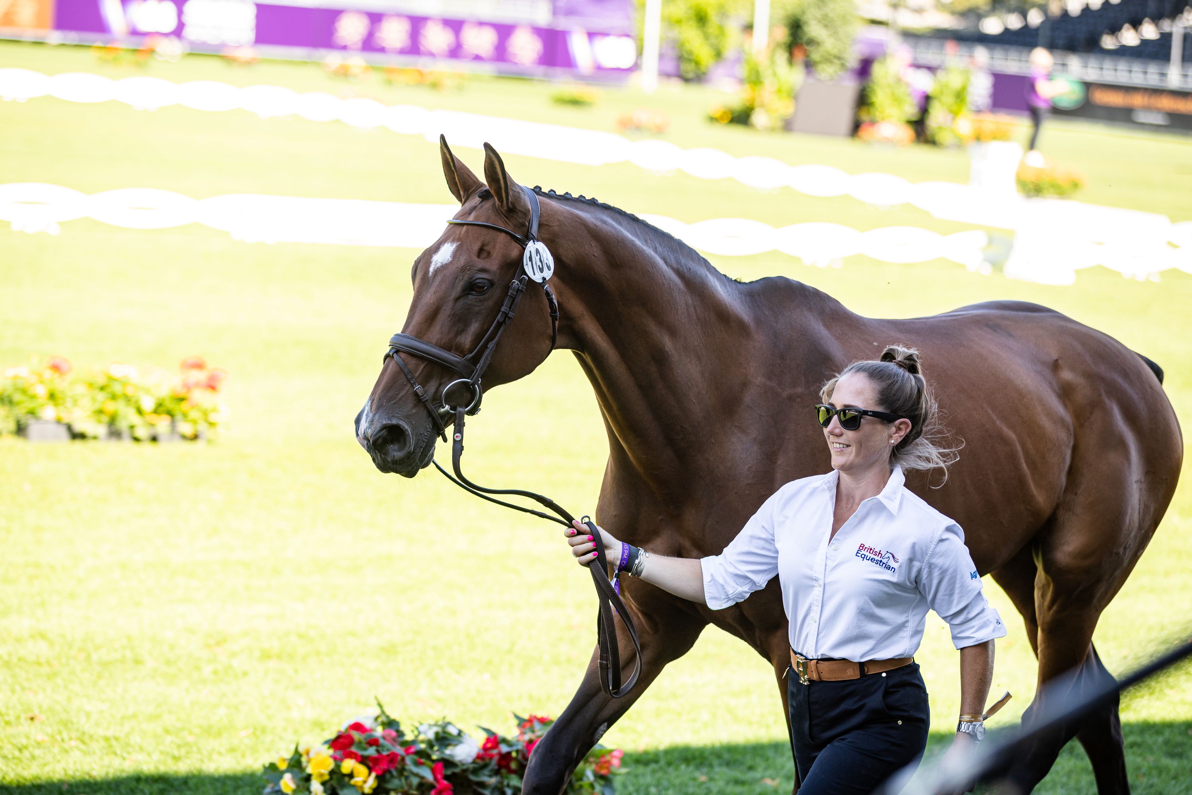 A rider in a white shirt bearing the ‘British Equestrian’ logo leads a bay horse, bearing start number 139, at a walk across the well-tended green lawn; in the foreground are colourful floral decorations, whilst in the background, the dressage arena’s boundary and the tournament backdrop are blurred.