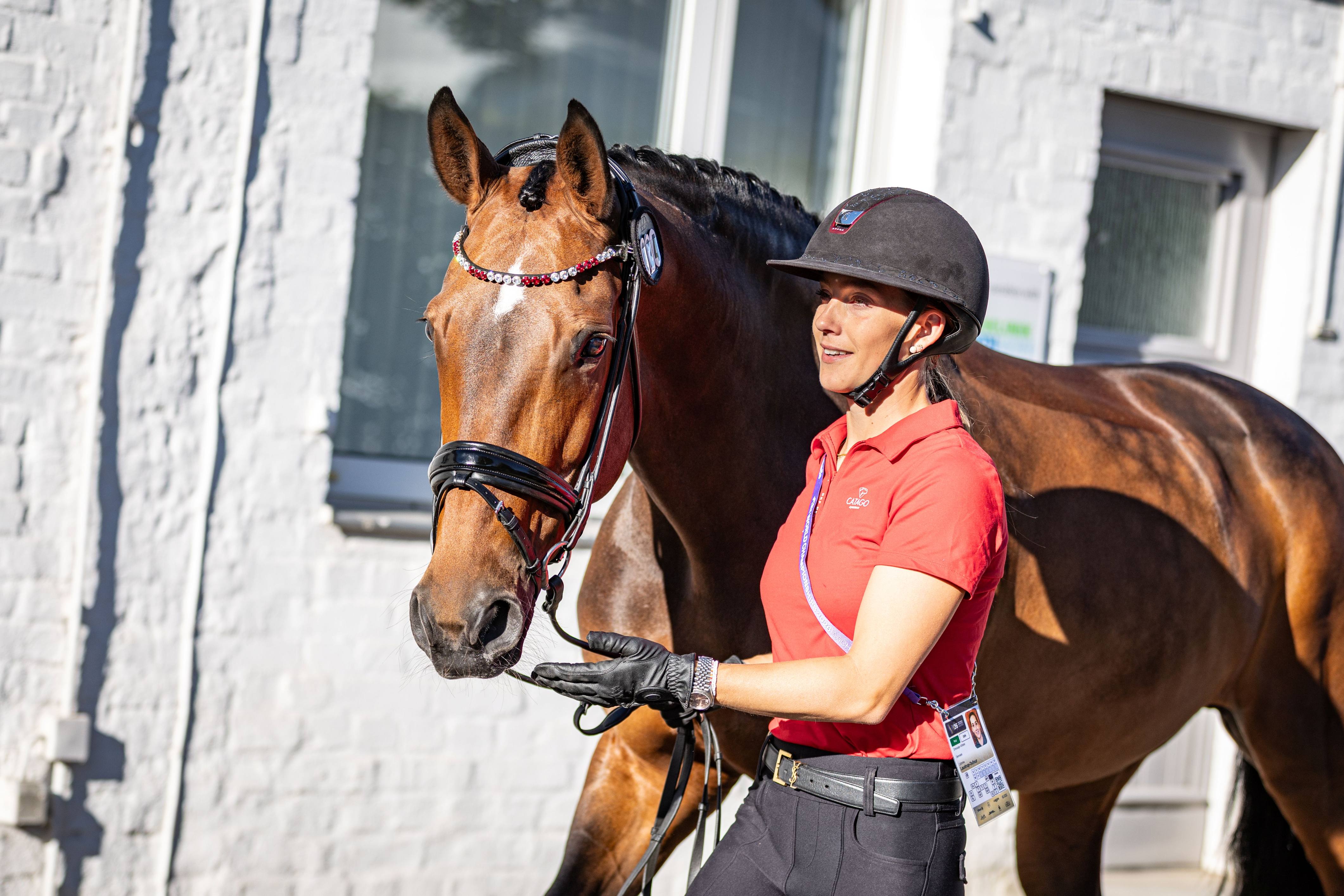 A woman in a red polo shirt and riding helmet is leading a brown horse by its bridle; the horse is wearing a browband adorned with red and white stones, and in the background a light-coloured house wall with windows can be seen.