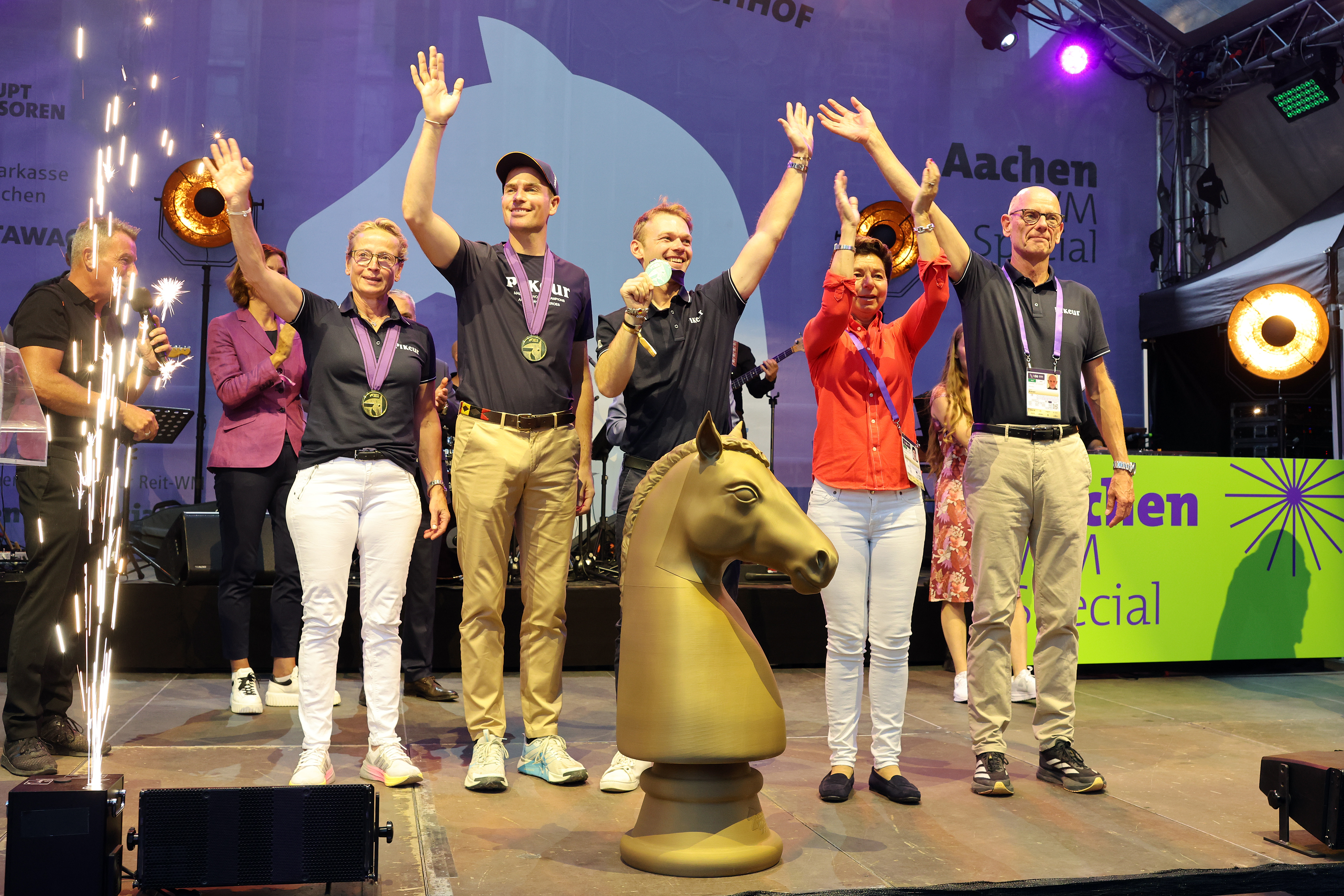 Gruppe von fünf geehrten Personen auf einer Bühne bei den FEI World Championships, alle mit Medaillen um den Hals, die lachend und winkend neben einer großen goldenen Pferdekopf Trophäe stehen; links sprühen Bühnenfontänen, im Hintergrund sind Musiker, Scheinwerfer und ein Banner mit der Aufschrift „Aachen“ zu sehen.