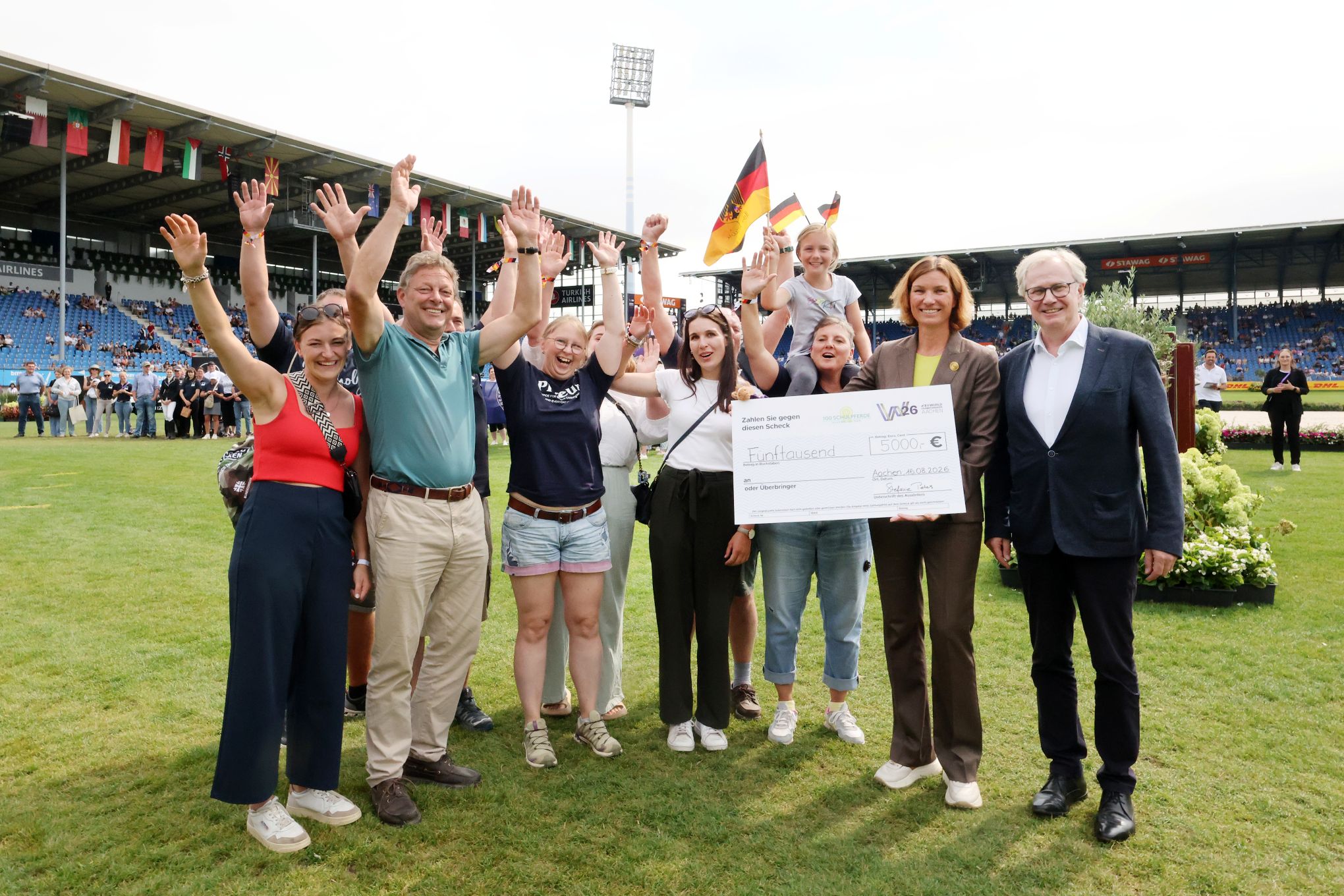 A group of cheerful people on the pitch at the Main Stadium, cheering and throwing their arms in the air as they hold aloft a large donation cheque for 5,000 euros; in the background, the spectator stands can be seen, decorated with national flags, whilst several German flags are visible on the pitch.