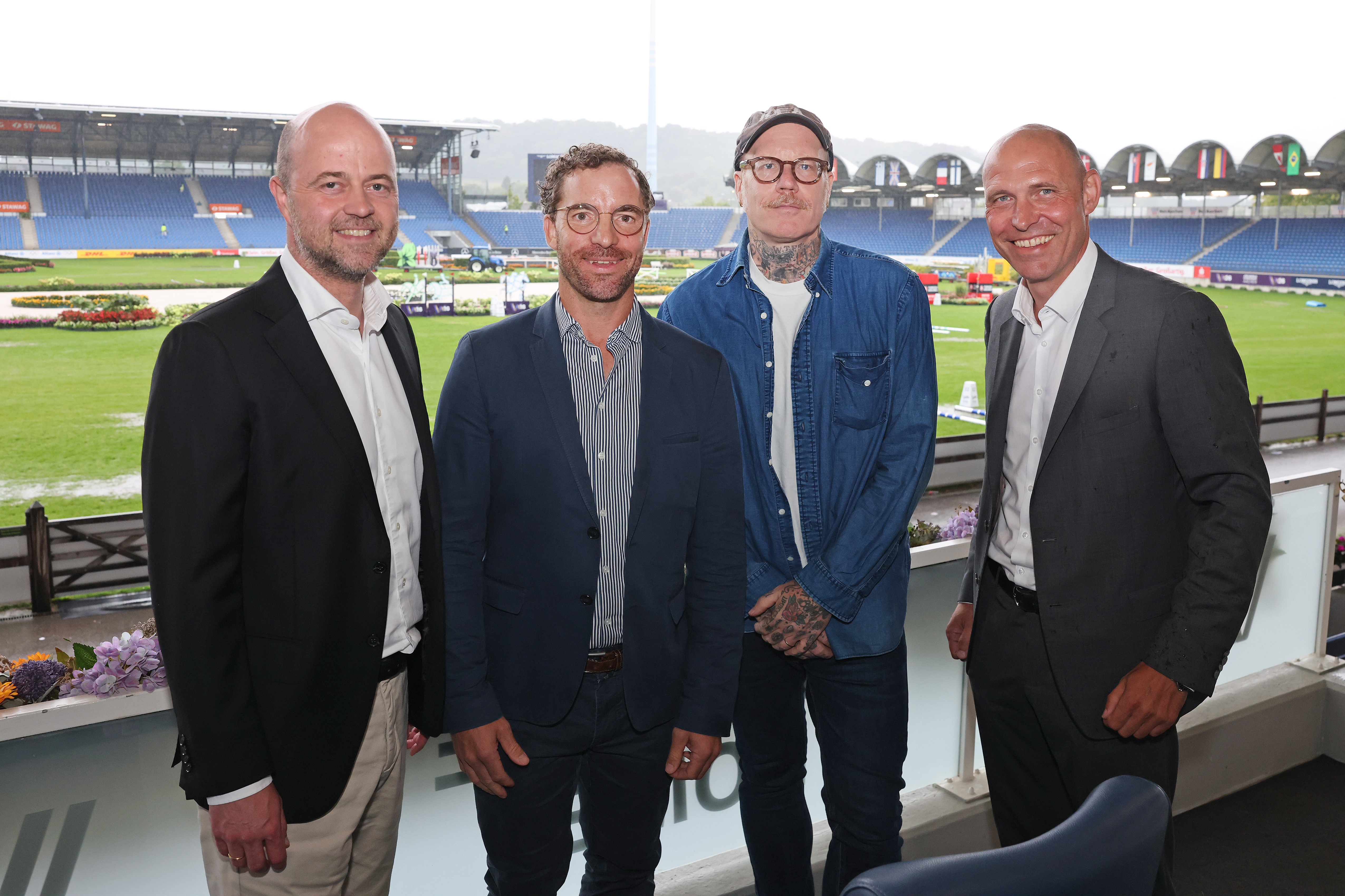 Vier Männer stehen für ein Gruppenfoto vor der Kulisse des Hauptstadions in der Aachener Soers mit Reitplatz, Tribünen und Länderflaggen. Drei tragen Anzüge, einer ein blaues Jeanshemd.