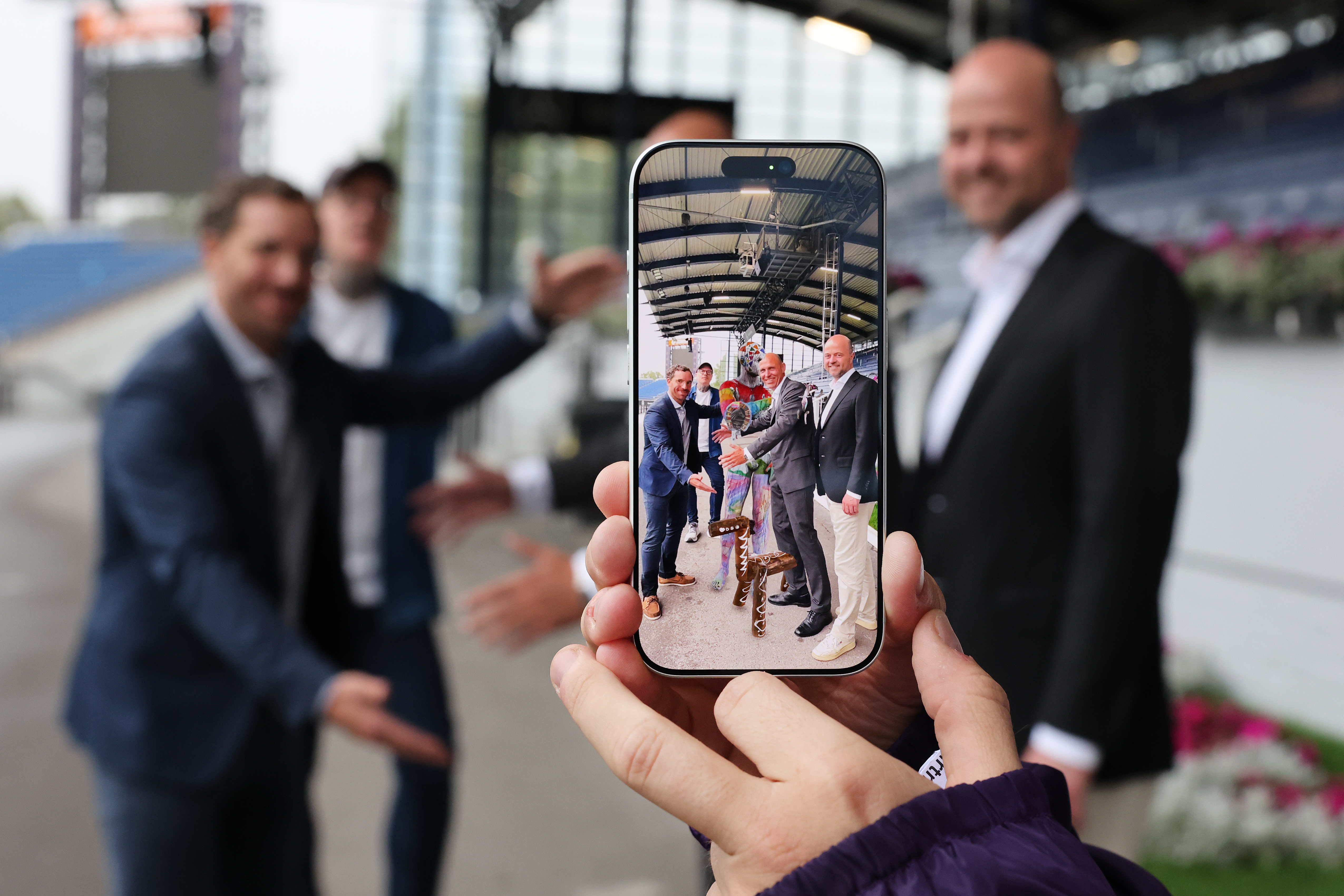 Eine Hand hält ein Smartphone, auf dessen Display vier Männer neben einer bunt bemalten Pferdefigur im Hauptstadion in der Aachener Soers posieren; im Hintergrund sind die Personen unscharf zu sehen.