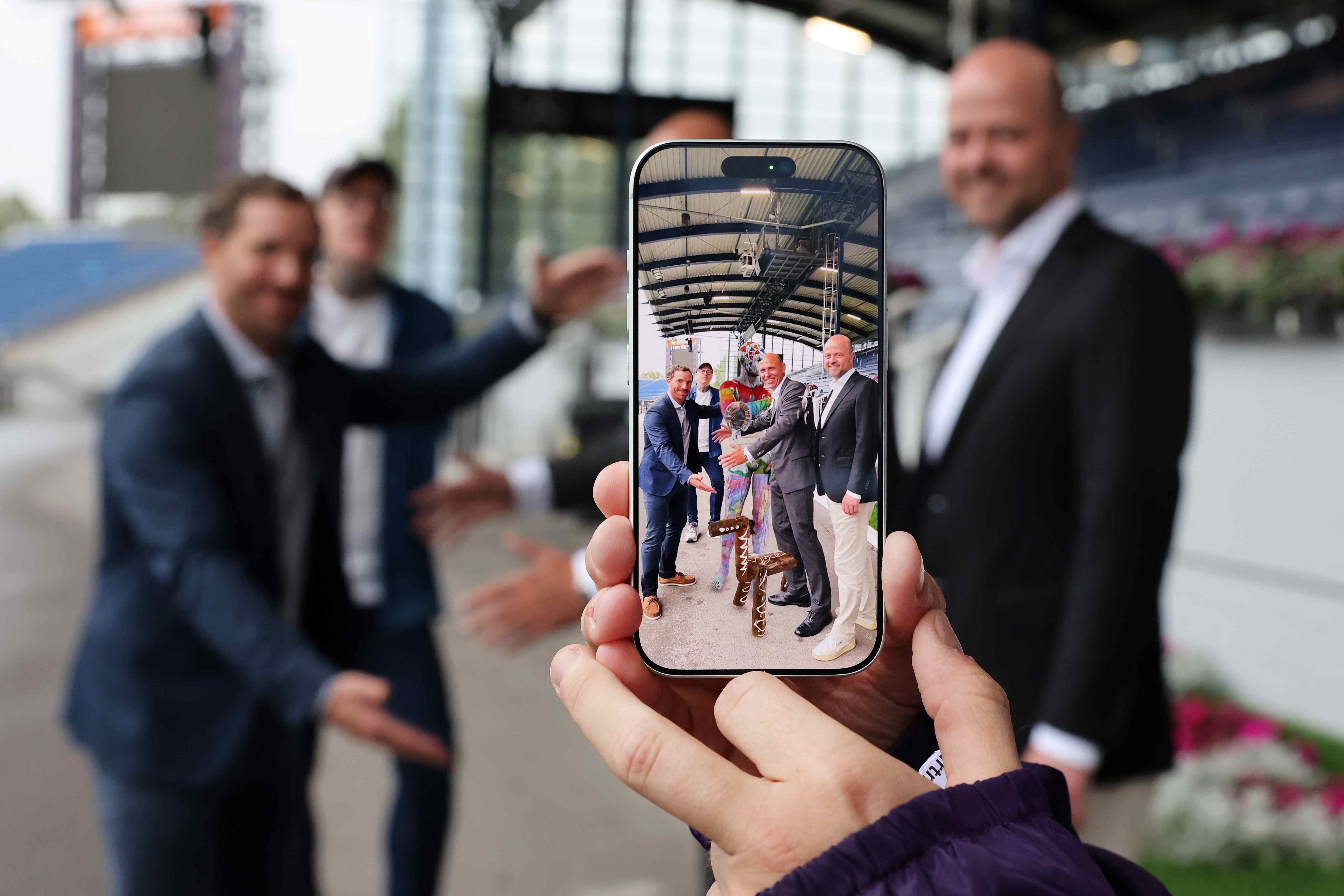 A hand is holding a smartphone, on whose screen four men are posing next to a colourfully painted horse figure in the Main Stadium at the Soers in Aachen; in the background, the figures are blurred.