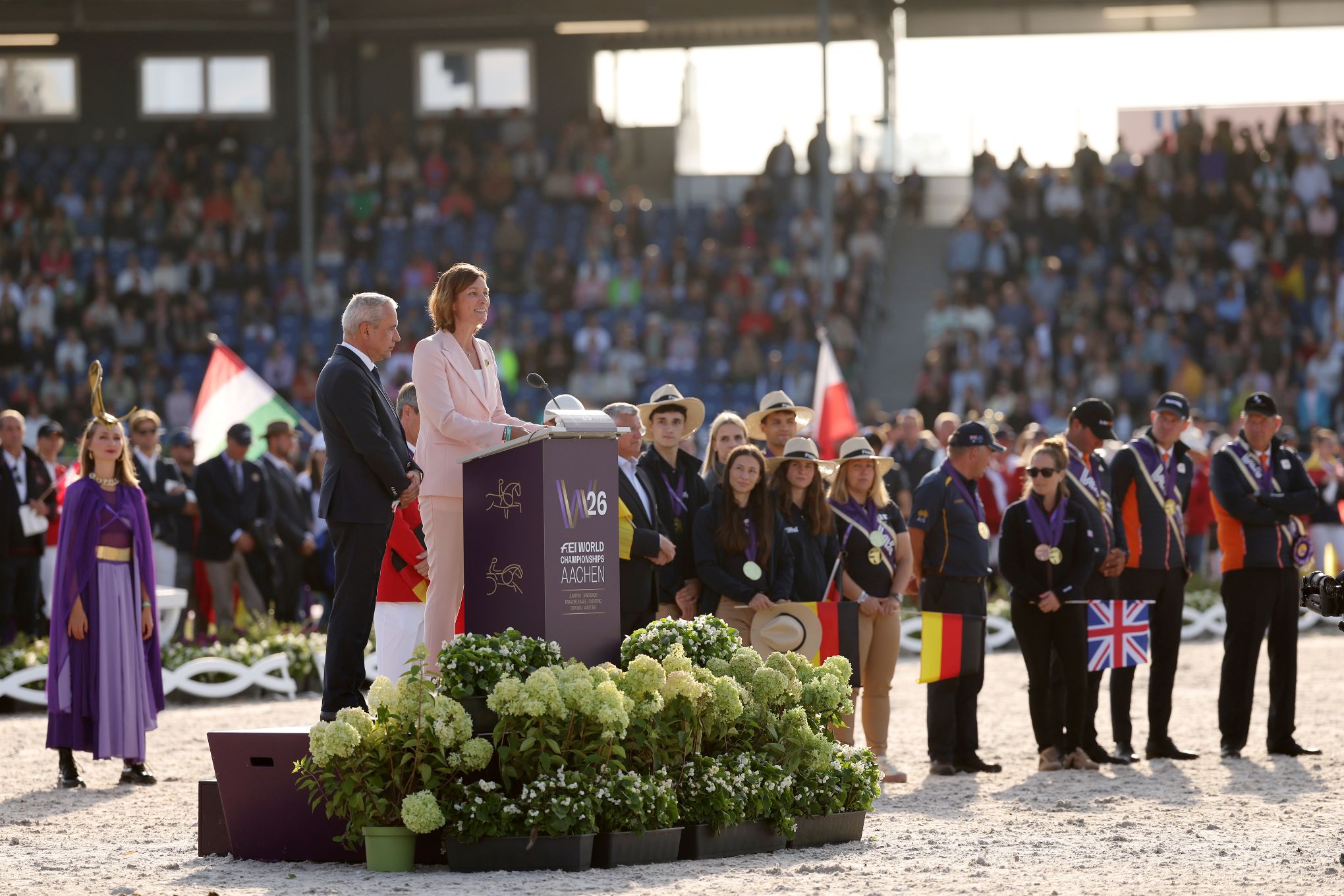 A woman in a pink trouser suit is speaking at a podium bearing the inscription ‘FEI World Championships Aachen 2026’ at an equestrian event. Standing next to her is a man in a dark suit. Behind them, participants wearing medals and holding international flags can be seen, along with numerous spectators; in the foreground, white flowers adorn the stage.