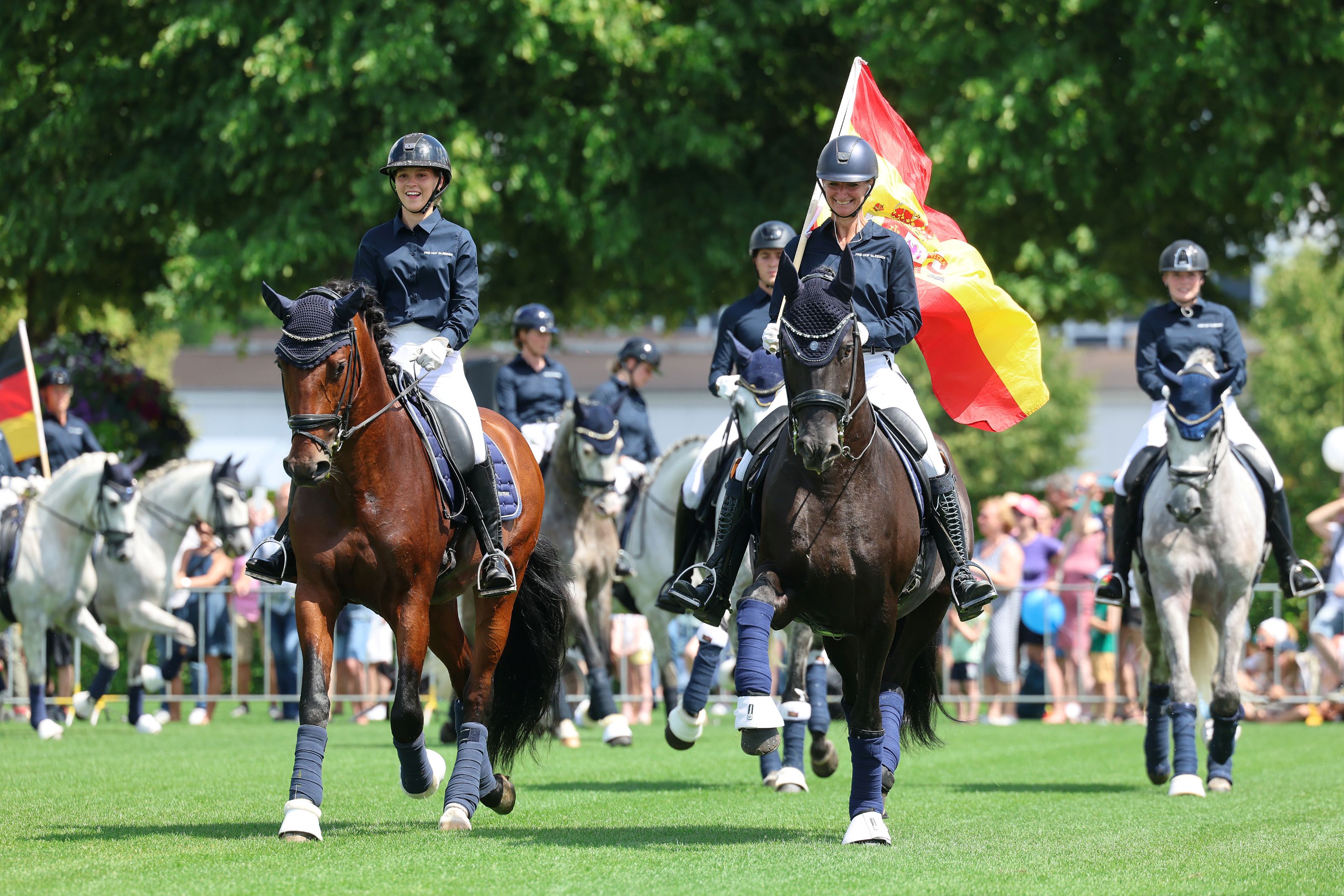 Riders in dark blue jackets are riding across the green lawn in the sunshine; in the foreground, a female rider on a dark horse is carrying a large red-yellow-red flag, whilst in the background there are more horses and a crowd of spectators.
