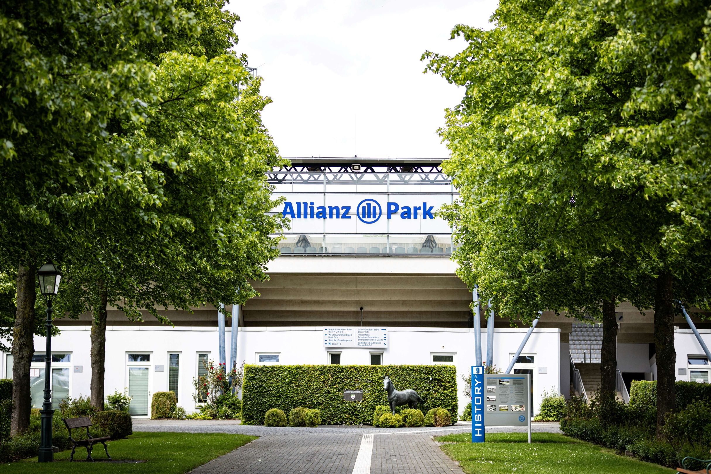A view through a tree-lined avenue towards the stand bearing the blue lettering ‘Allianz Park’; in the foreground are a path, an information board and a small sculpture of a horse in front of the stadium building.
