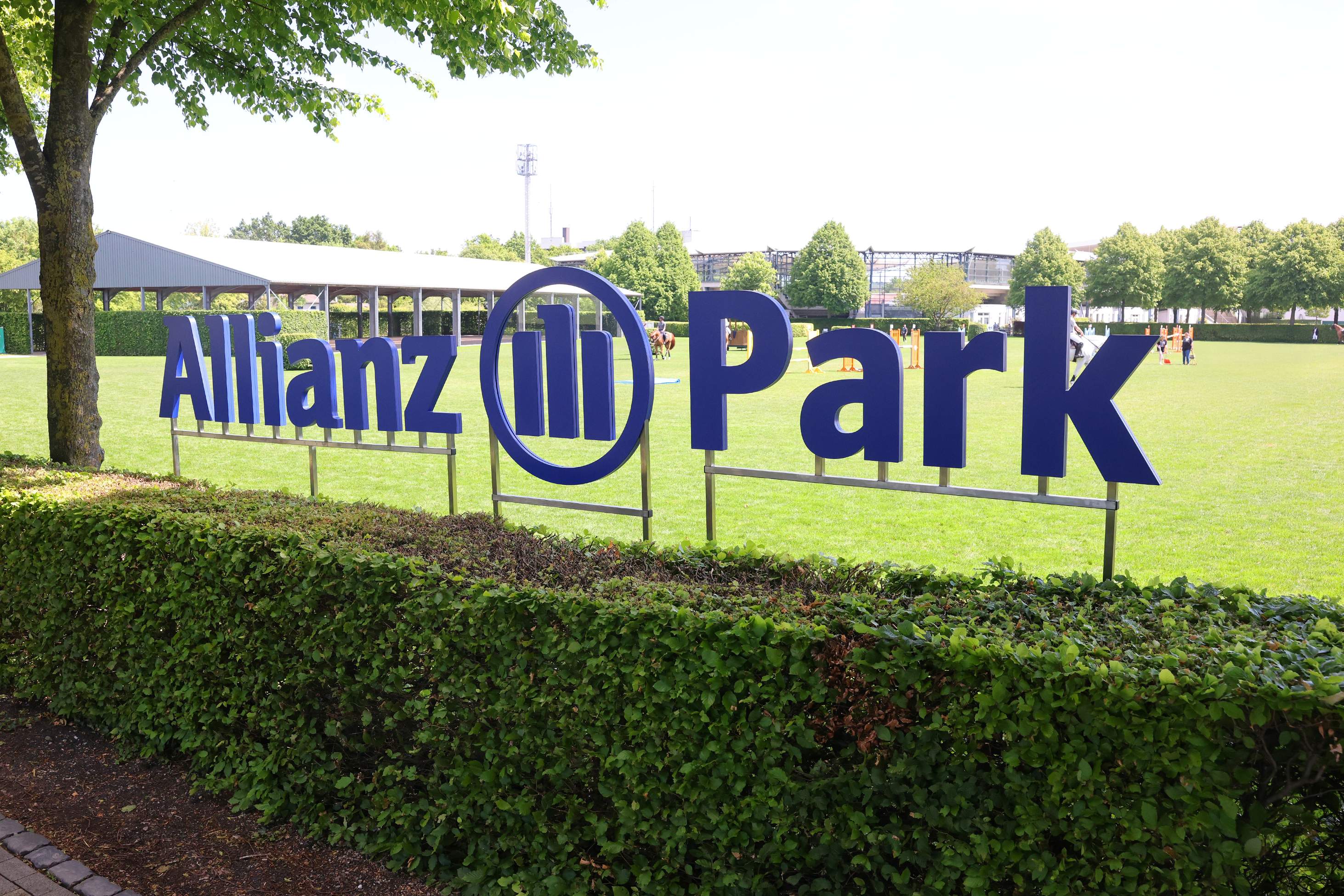 A large blue sign reading ‘Allianz Park’ stands at the edge of a green riding arena; in the background, show jumping obstacles and riders can be seen, framed by trees and hedges.