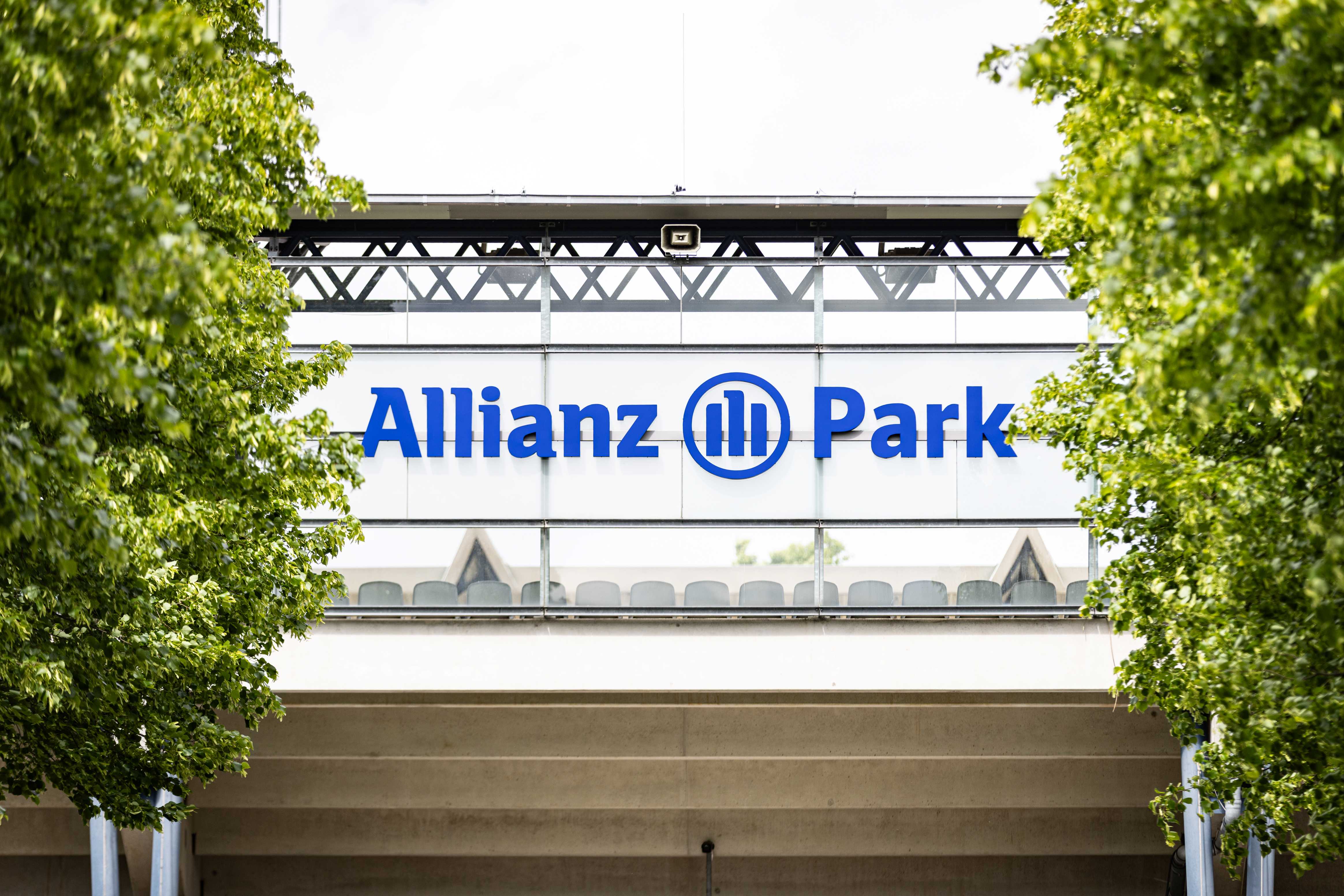 A close-up of a glass façade featuring the blue lettering ‘Allianz Park’ and the Allianz logo, framed by green trees.
