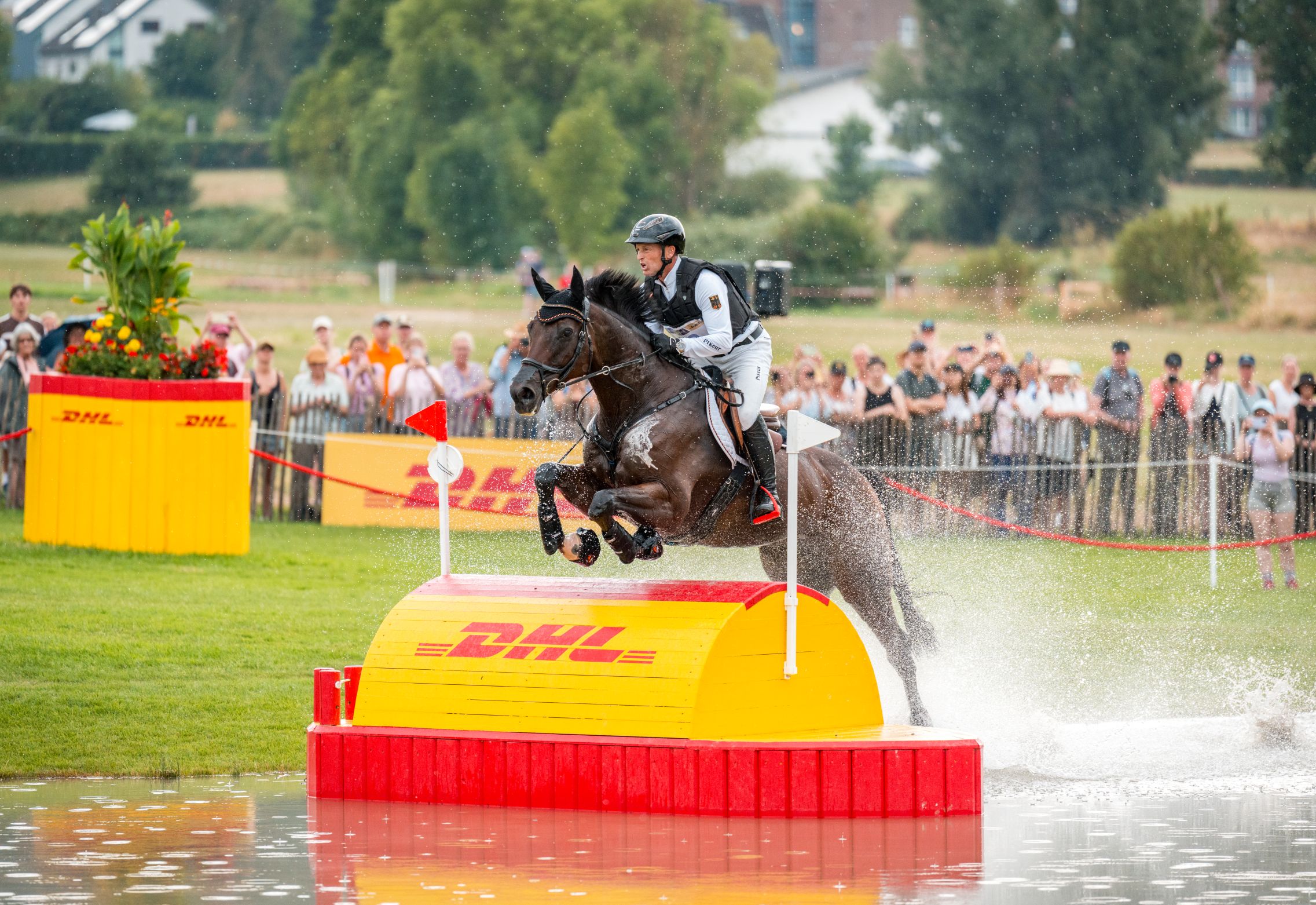 An eventer jumps over a yellow-and-red DHL obstacle in a water jump on a bay horse, sending water spraying high into the air; in the background, numerous spectators are standing at the edge of the meadow watching the ride.
