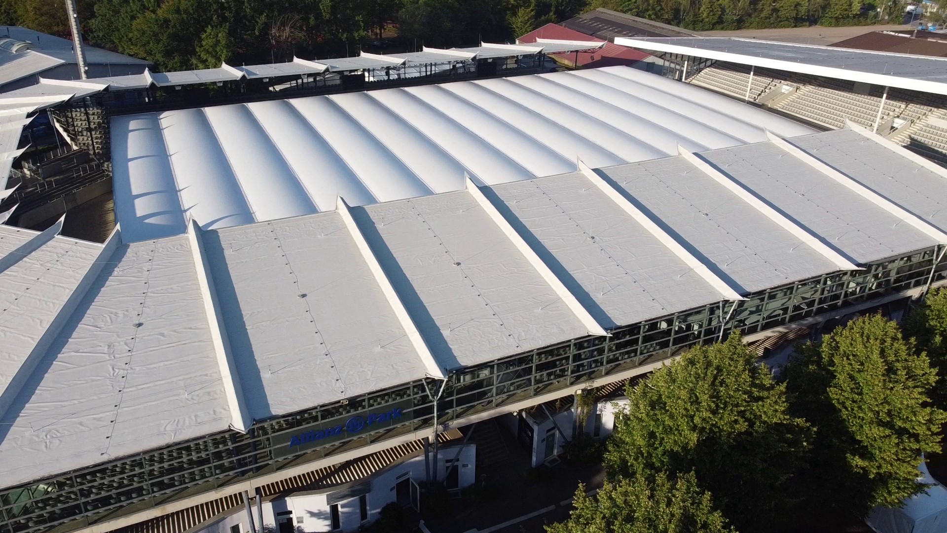 Aerial view of a stadium at the Allianz Park featuring a large, white membrane roof: several parallel roof sections span the stands and the interior; trees can be seen in the foreground, with further halls and facilities visible in the background.