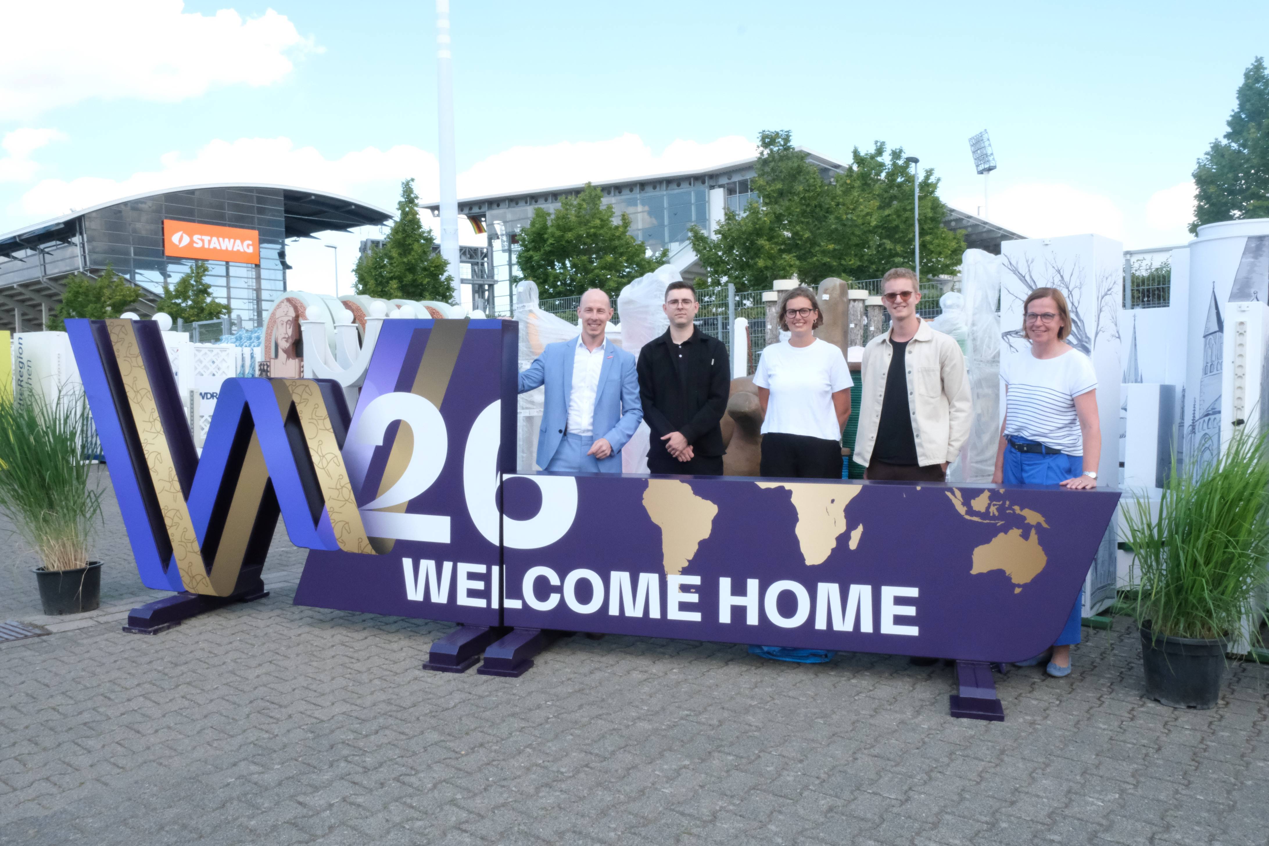 A group of five people is standing in the outdoor area behind a large, purple-and-gold sculpture bearing the inscription ‘W26 WELCOME HOME’ and stylised maps of the world; in the background, trees, a stadium building bearing the ‘STAWAG’ logo and other decorative elements can be seen.