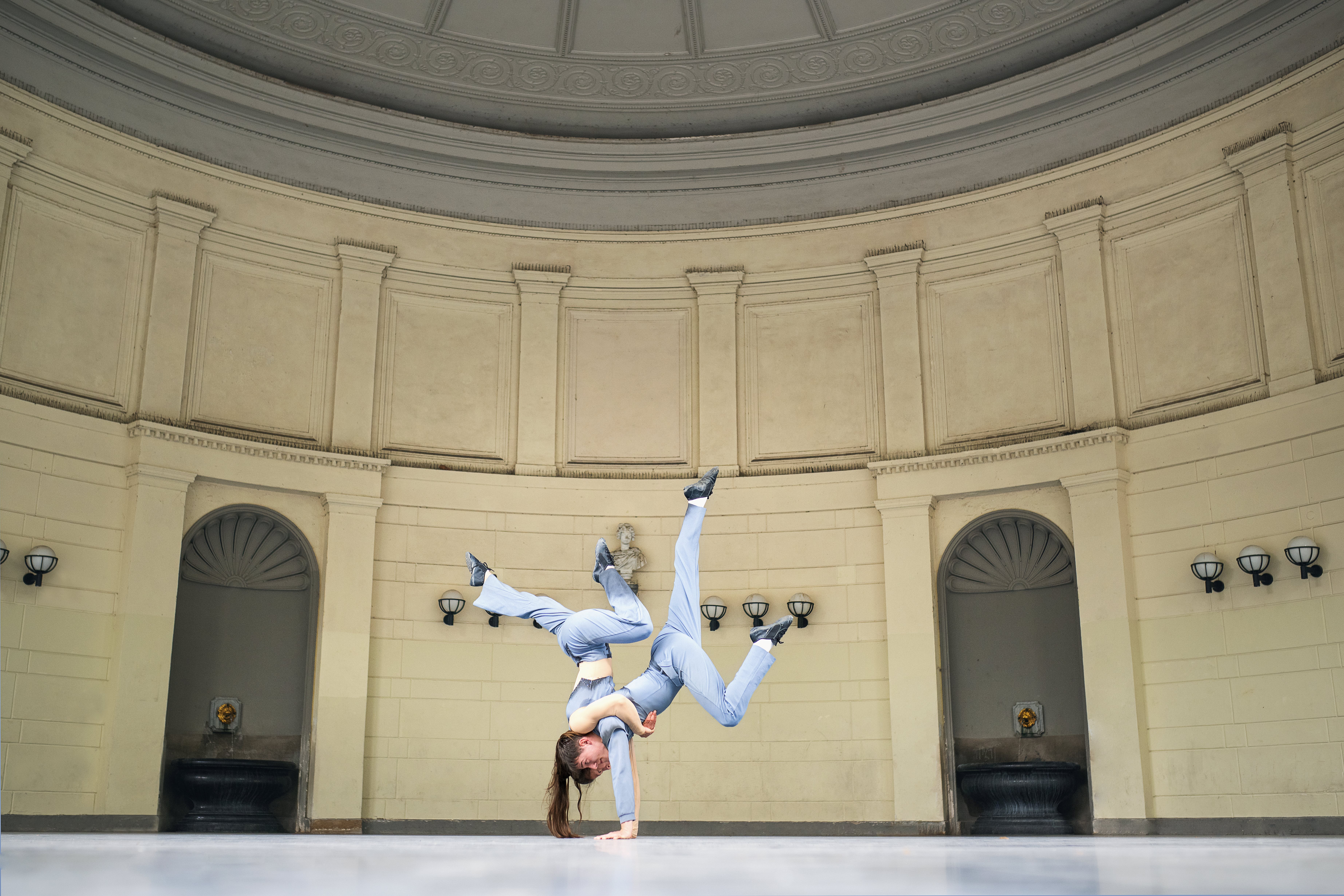 Two acrobats dressed in light blue are performing a complex partner manoeuvre in a historic rotunda, in which one athlete, in a handstand, balances the other on her back whilst both legs are dynamically stretched out into the air.