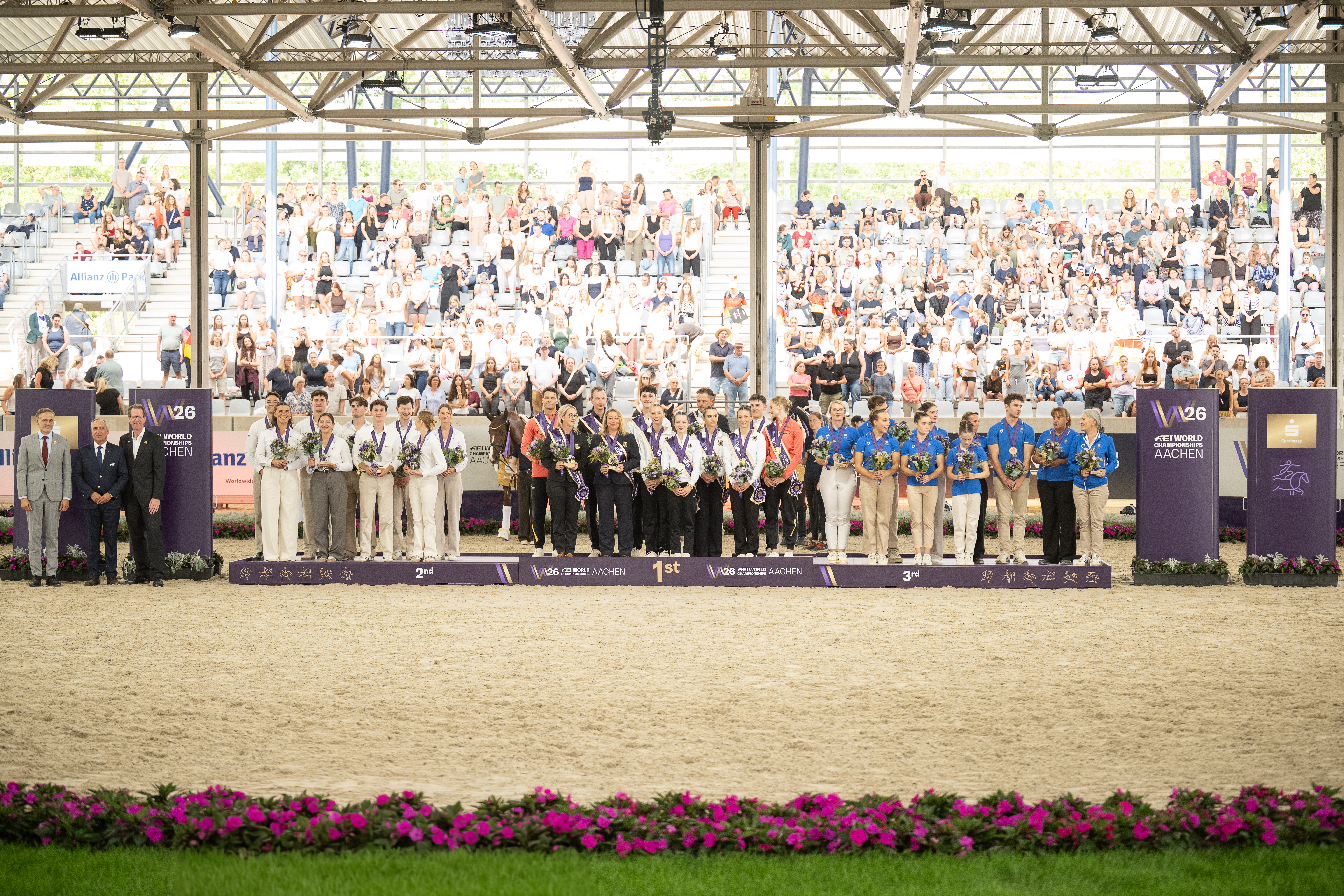 Siegerehrung bei den FEI World Championships Aachen: Auf einem breiten Podest mit den Aufschriften „2nd“, „1st“ und „3rd“ stehen mehrere Voltigier Teams in Wettkampfbekleidung mit Medaillen und Blumensträußen, rechts und links flankiert von Offiziellen. Im Hintergrund füllt ein großes, überdachtes Tribünenrund mit zahlreichen Zuschauern das Bild; vorne säumt ein Beet mit pink blühenden Blumen den Sandplatz.