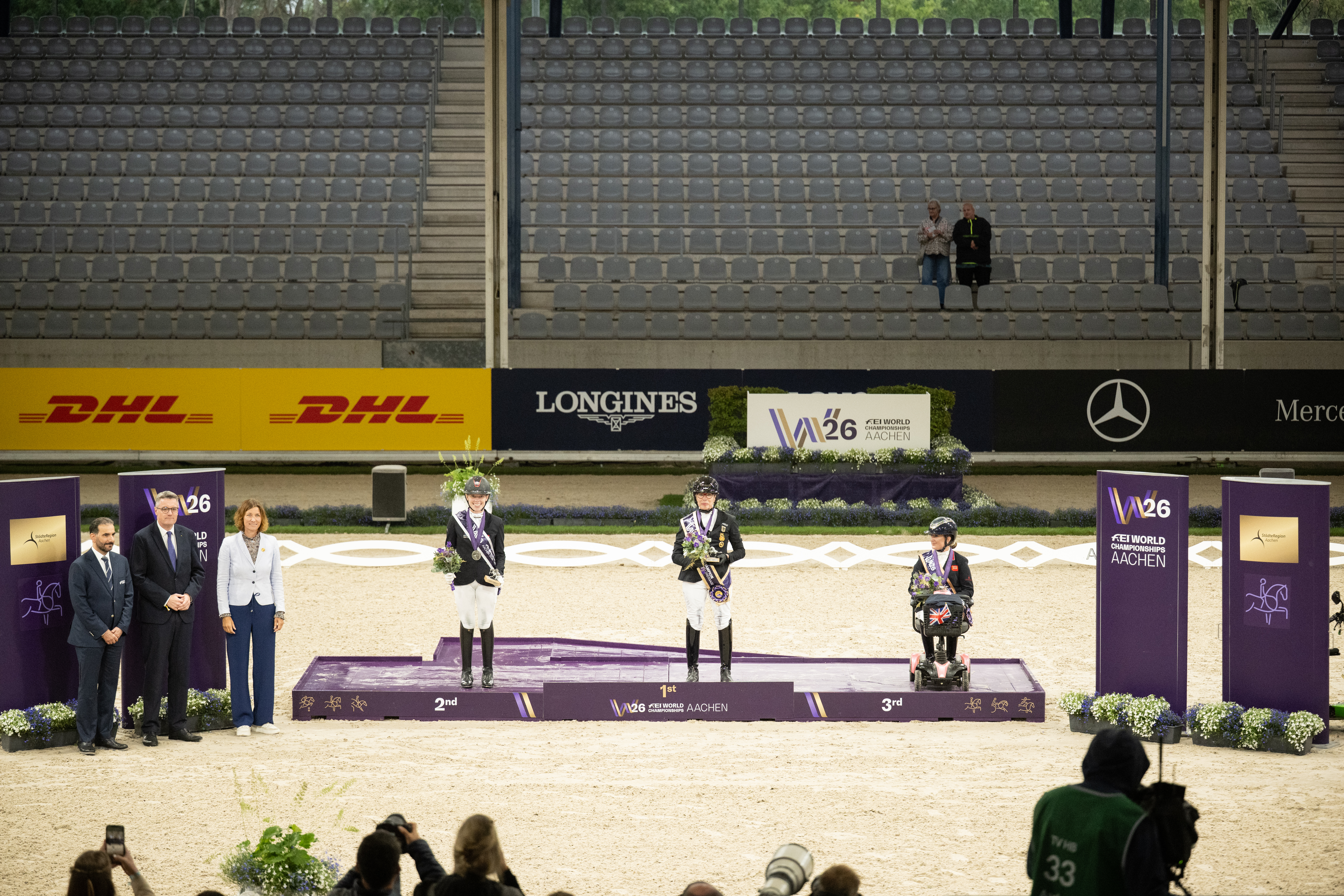 Siegerehrung im Stadion 3: Drei ausgezeichnete Reiterinnen und Reiter stehen auf dem Podium auf den Plätzen eins bis drei, flankiert von vier Ehrengästen. Im Hintergrund sind Tribünen, Sponsorenlogos und das Logo der FEI World Championships Aachen 2026 zu sehen.