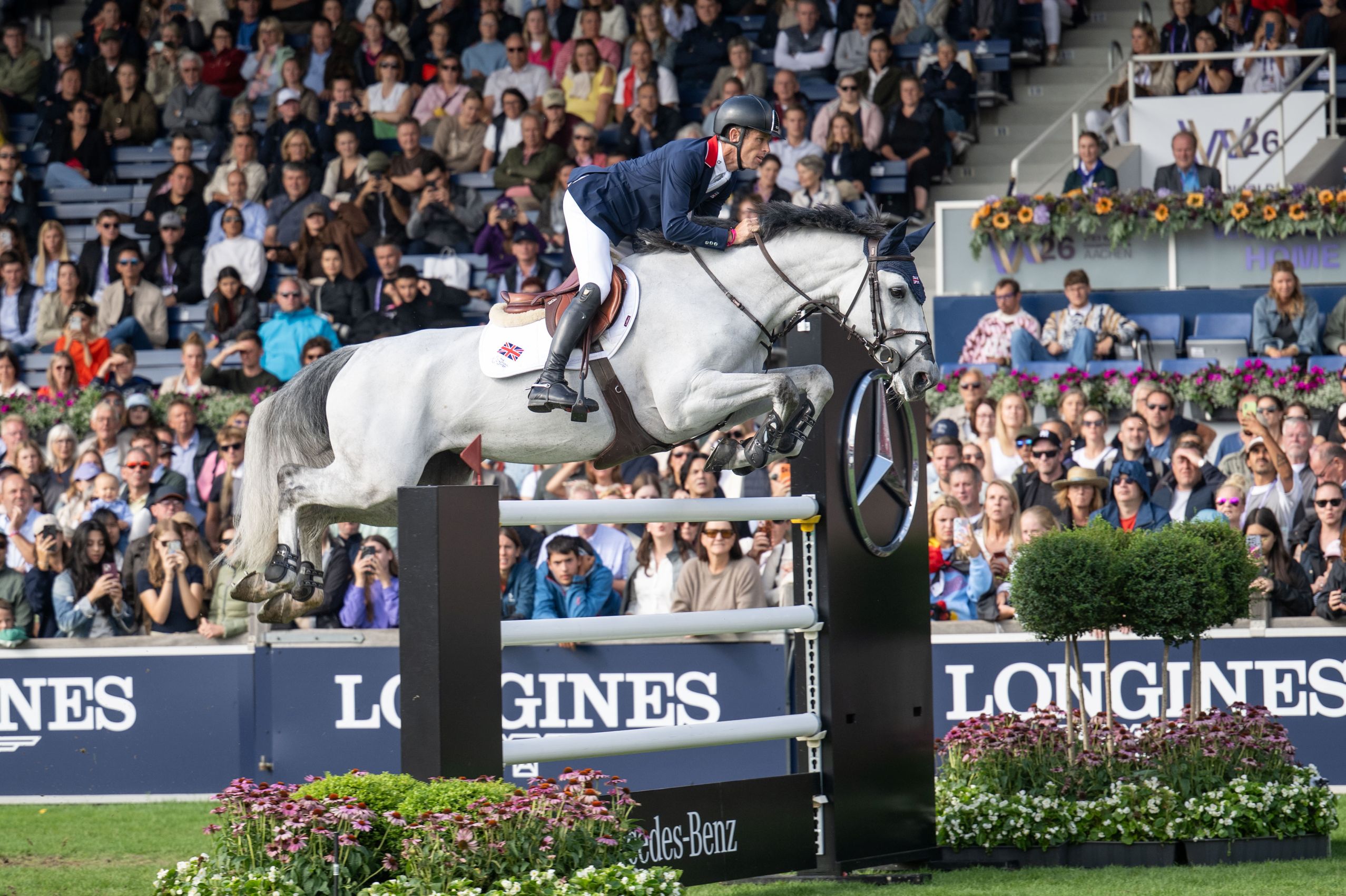 A rider in a dark blue jacket and white riding breeches jumps over an obstacle on a grey horse. A British flag can be seen on the saddlecloth. In the background, a large crowd of spectators are watching the show jumping competition.