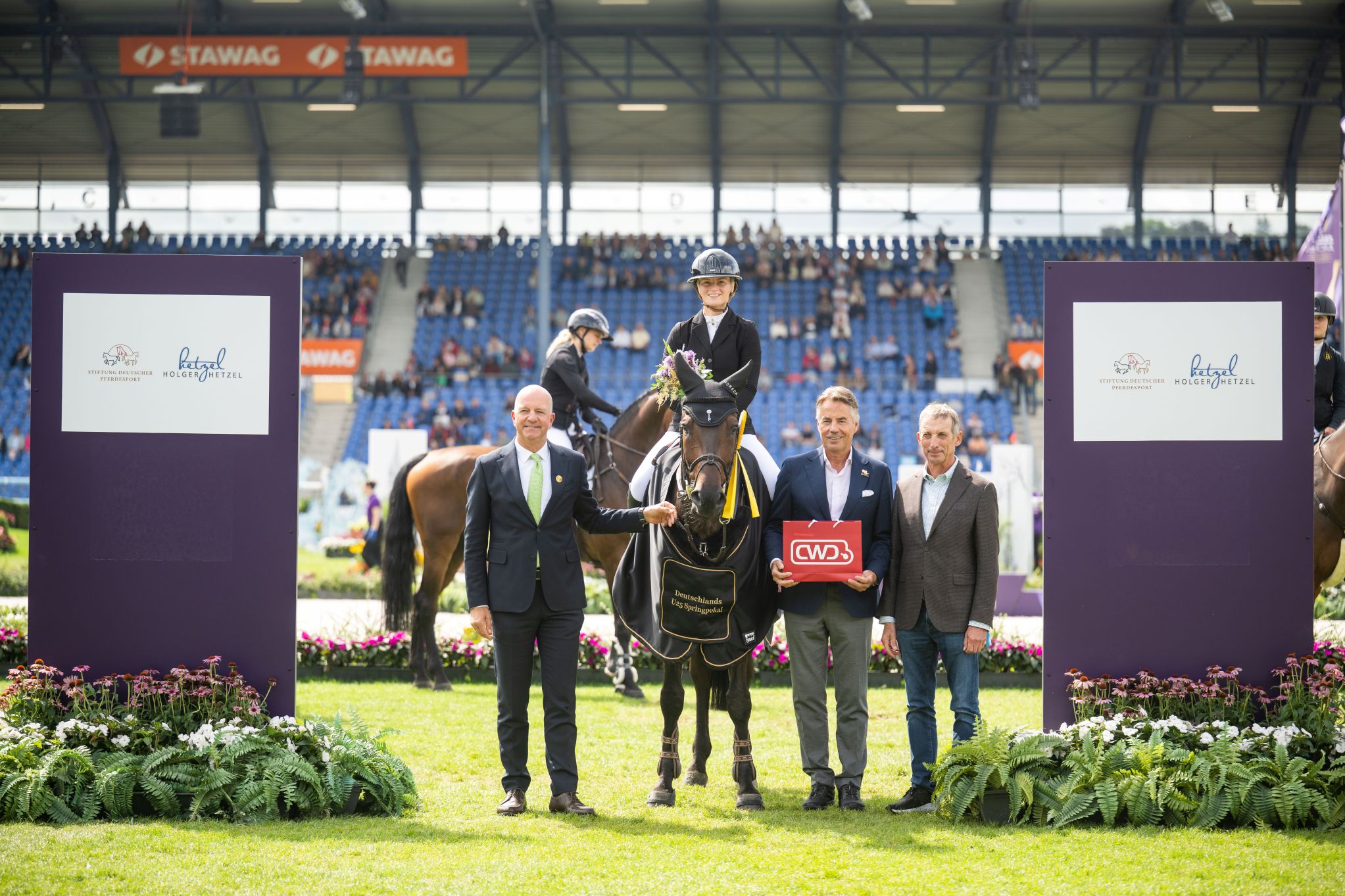 Prize-giving ceremony at the Main Stadium: A female rider is sitting on a horse with a black blanket; three men are standing in front of her for a photograph; one is holding a red CWD bag; in the background, the stands are full and other riders can be seen.