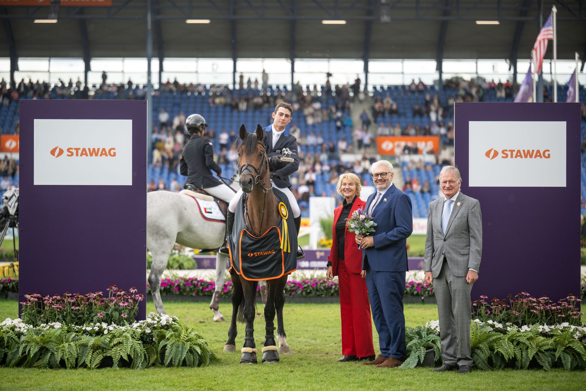 A prize-giving ceremony in a packed arena: a rider is sitting on a bay horse with a yellow bow and a STAWAG blanket. Standing next to him are a woman in red and two men holding a bouquet of flowers; in the background, another rider can be seen on a grey horse. Large STAWAG advertising hoardings stand on either side.