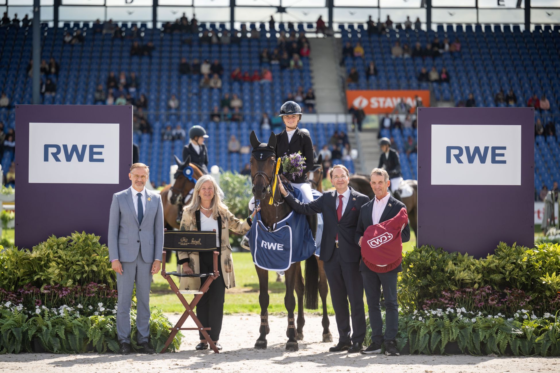 Prize-giving ceremony at the Main Stadium: A rider is sitting on a horse with a blue RWE blanket, holding a bouquet of flowers, with four officials standing next to her for a photograph; on the right, someone is holding a red CWD bag; in the background, more riders and the stands can be seen.