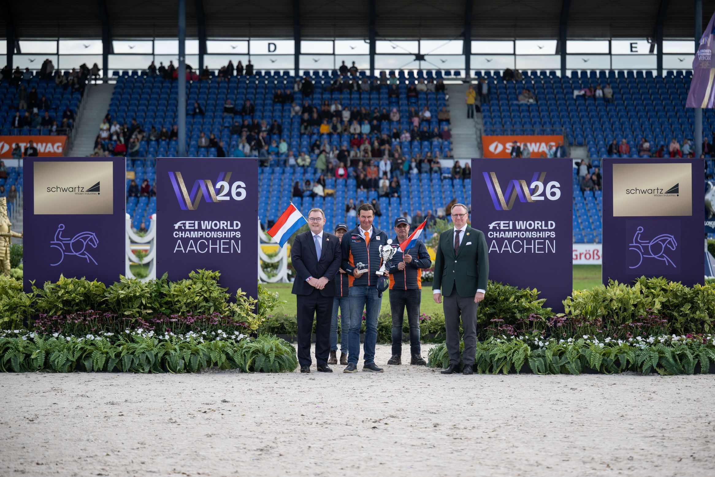 Prize-giving ceremony at the stadium: Five men are standing next to large ‘W26 FEI World Championships Aachen’ signs and ‘schwartz’ sponsor boards; one is holding the trophy, his team are holding Dutch flags, and the stands can be seen in the background.