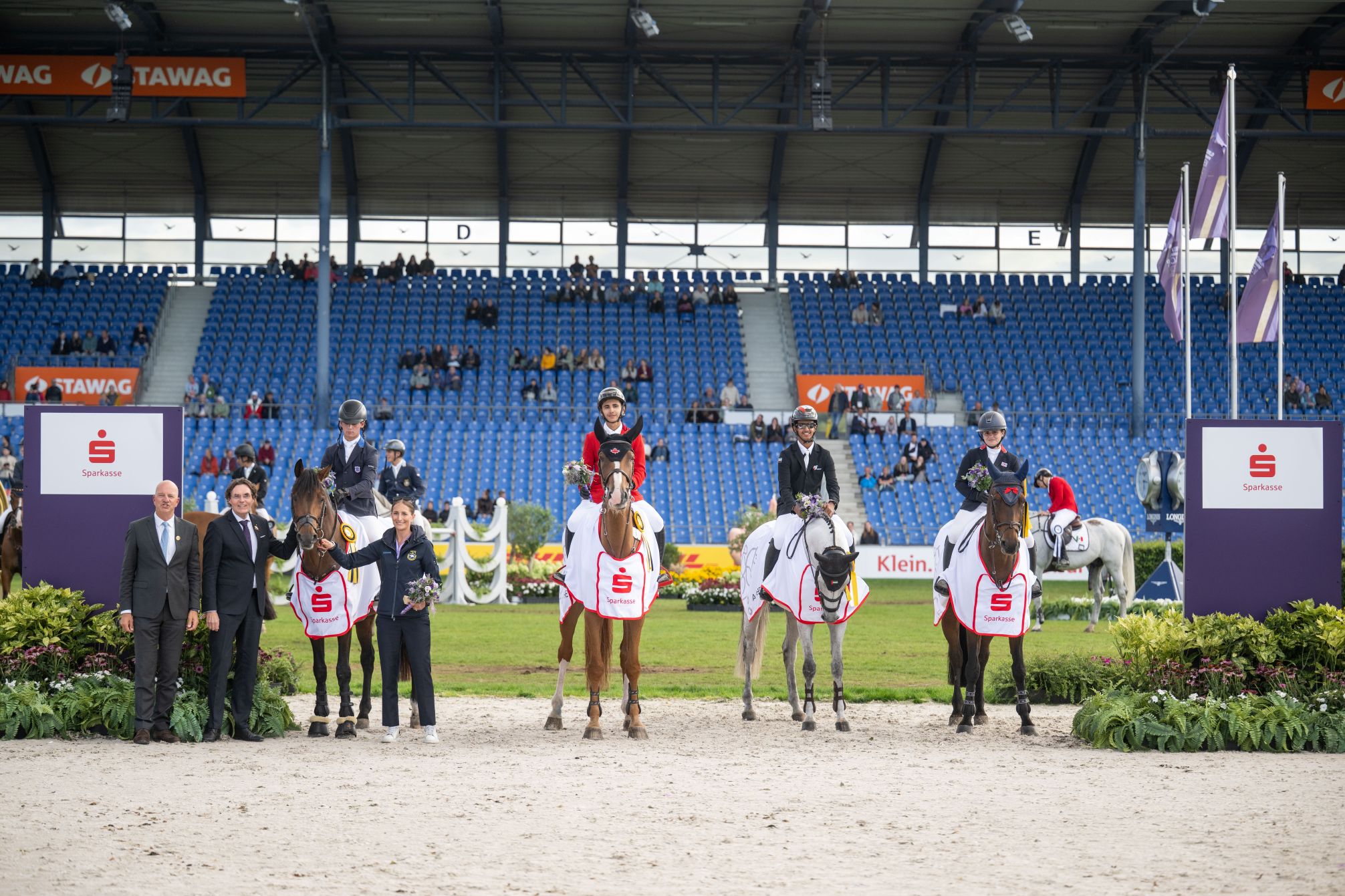 Equestrian prize-giving ceremony: Four riders are seated on their horses, which are draped in white Sparkasse blankets, and are holding bouquets of flowers. Three guests of honour are standing beside them; in the background, further horses, grandstands and event banners can be seen.