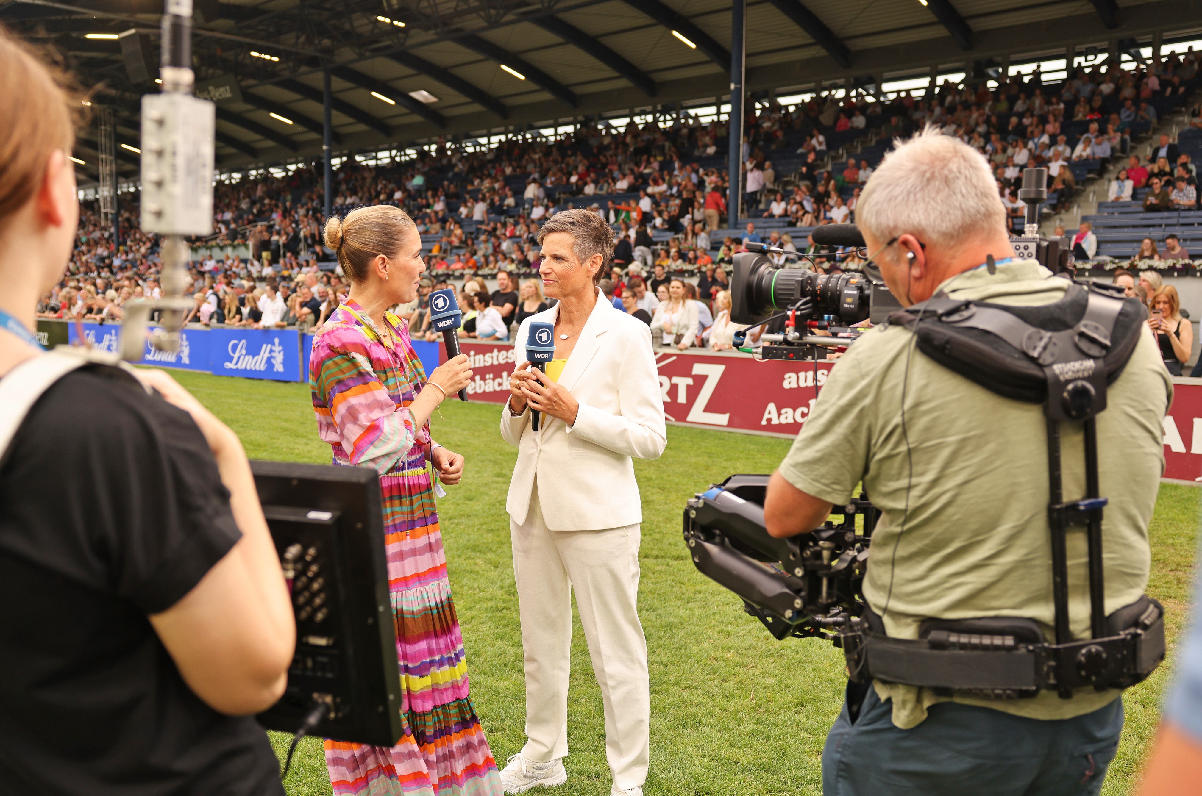 Two TV hosts are standing on the grass of a packed equestrian stadium conducting an interview, whilst in the foreground a cameraman with a Steadicam and an assistant with a monitor are filming; in the background, spectator stands and advertising boards can be seen.
